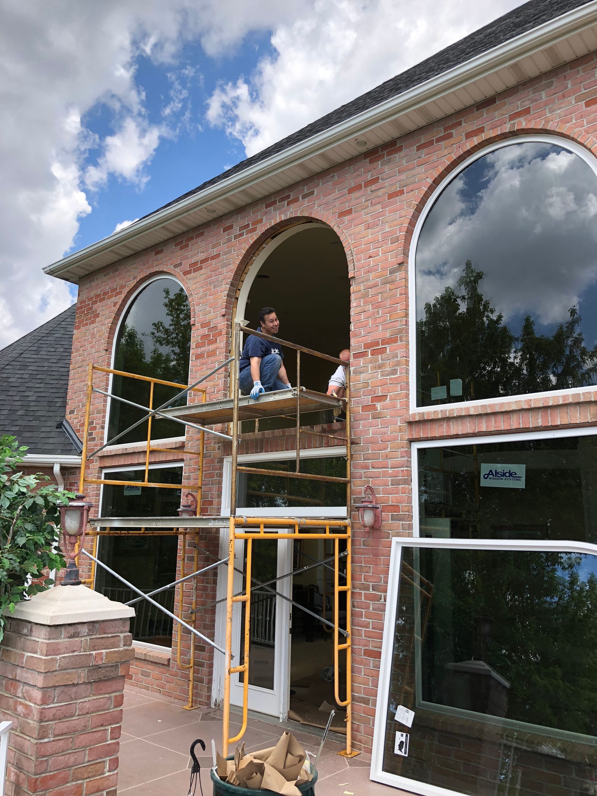 Construction workers replace windows on a brick house, using scaffolding on a sunny day.