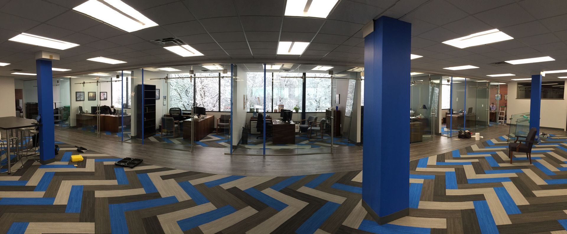Office interior with glass walls, desks, blue and gray zigzag patterned carpet, and blue support columns.