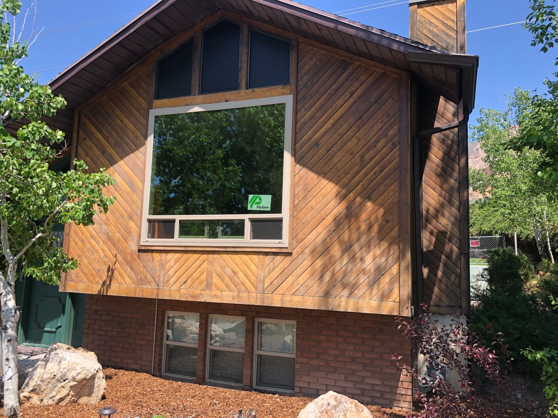 Brown wooden house with large central window and brick foundation, surrounded by trees.