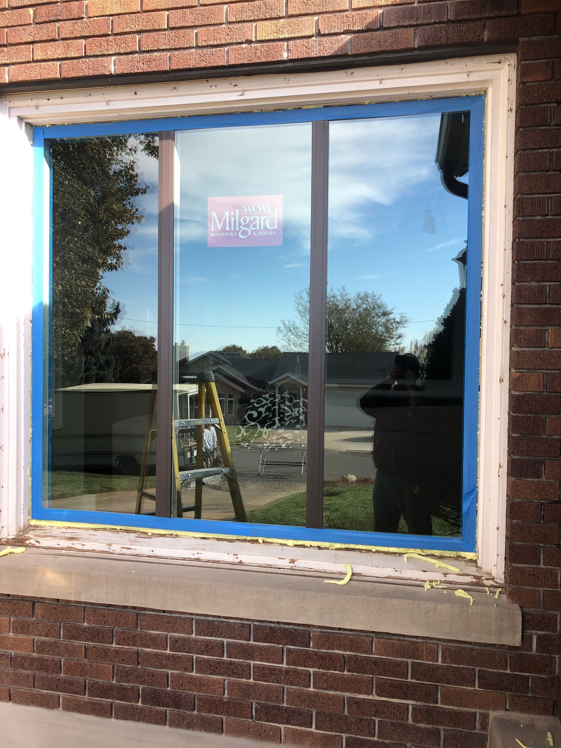 Window installation with blue tape on a brick house, reflecting sky and a ladder.