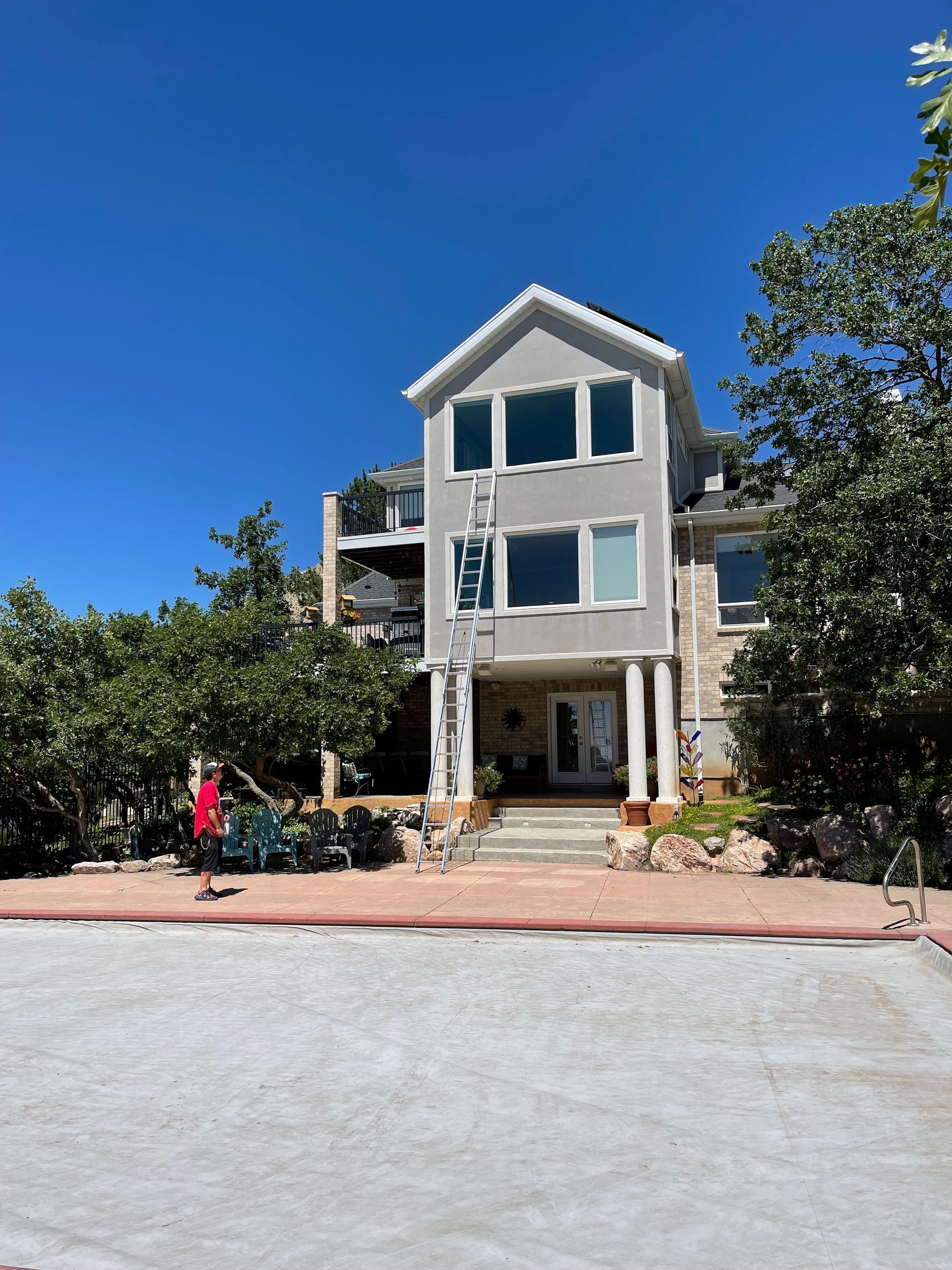 Three-story house with light grey siding and a white door, surrounded by trees, under a bright blue sky.