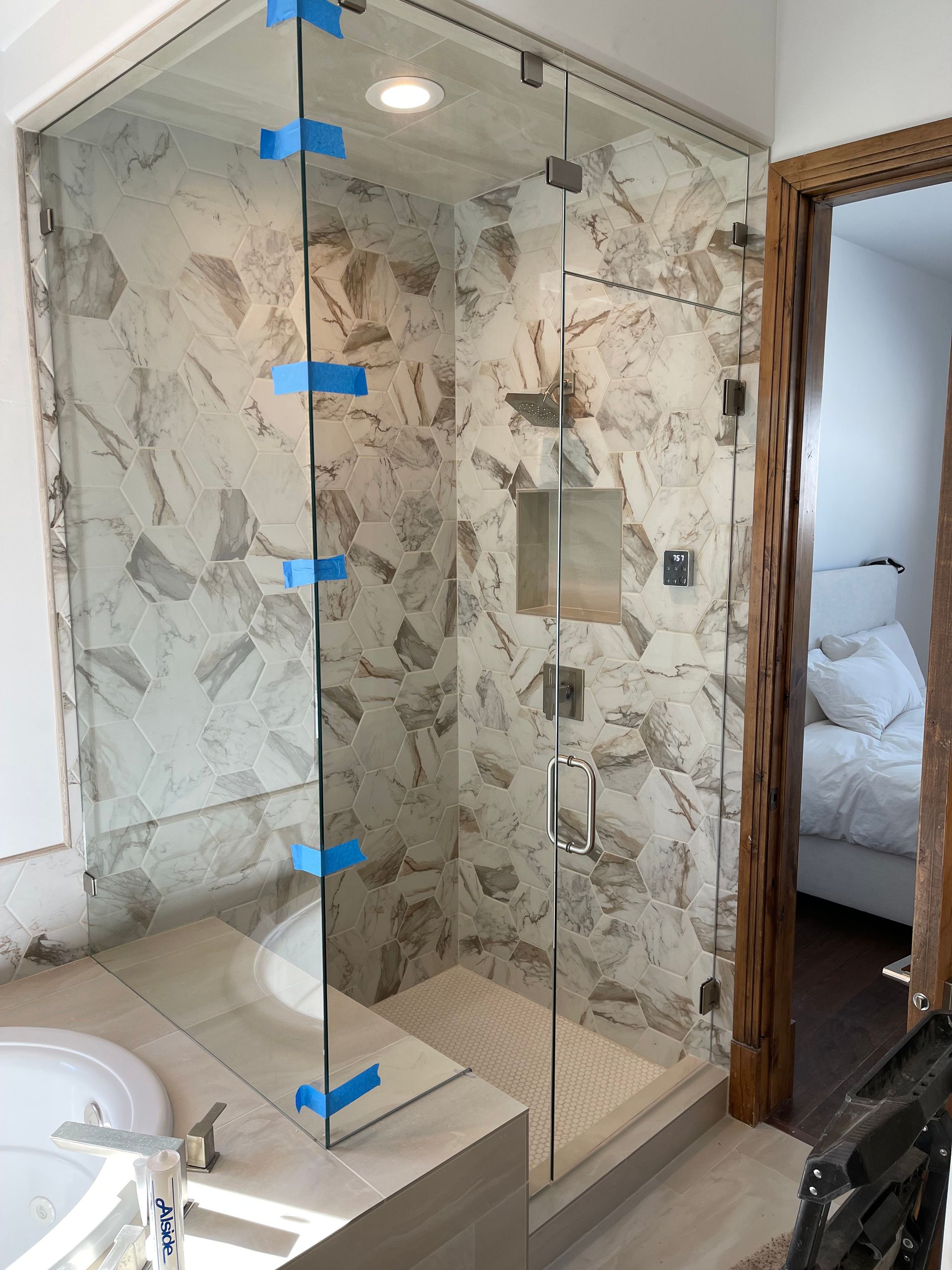 Glass shower enclosure with marble-patterned tile walls, door open to a bedroom.