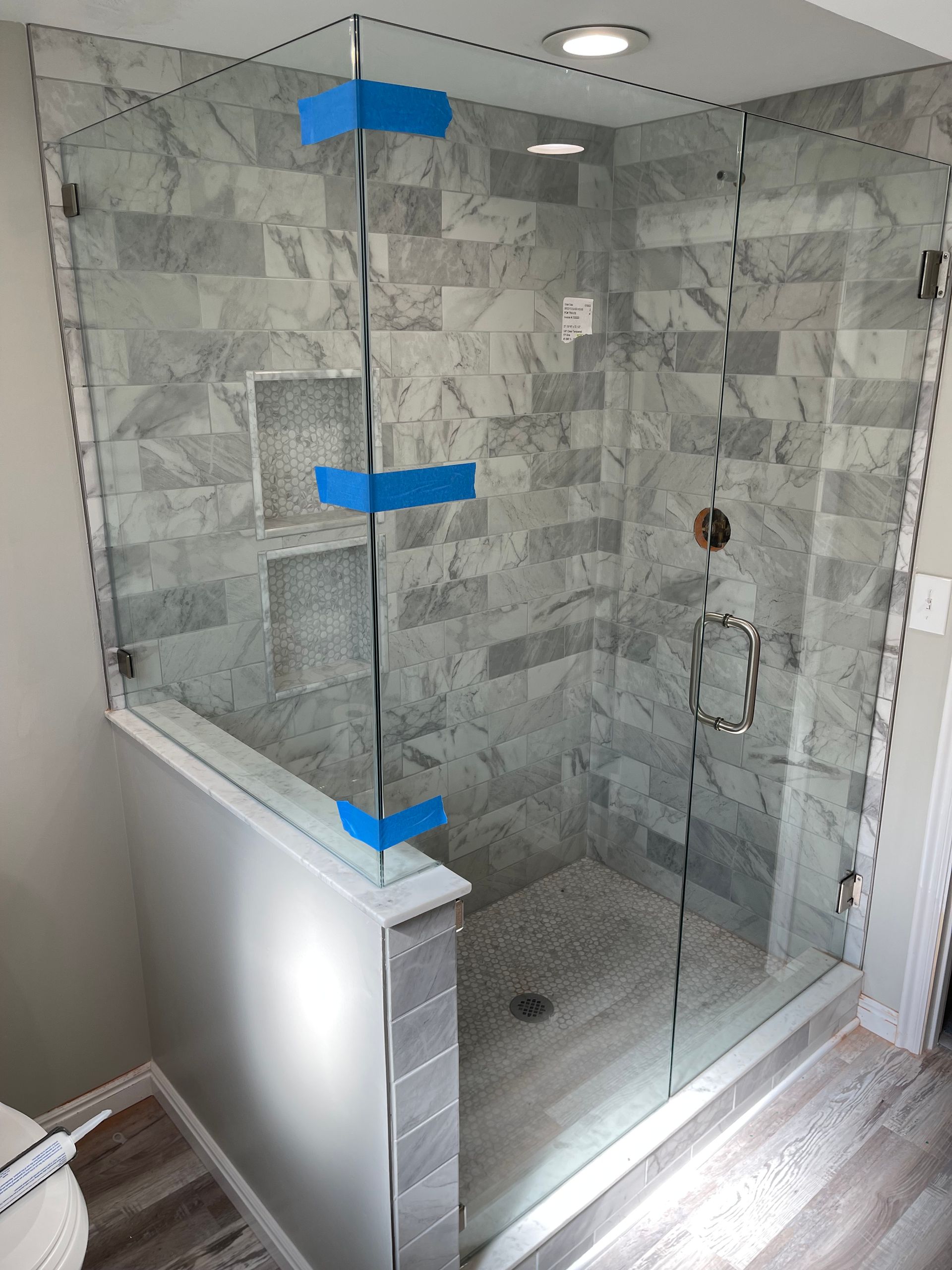 Glass shower enclosure with gray tiled walls and floor; blue tape marks where glass panels meet.