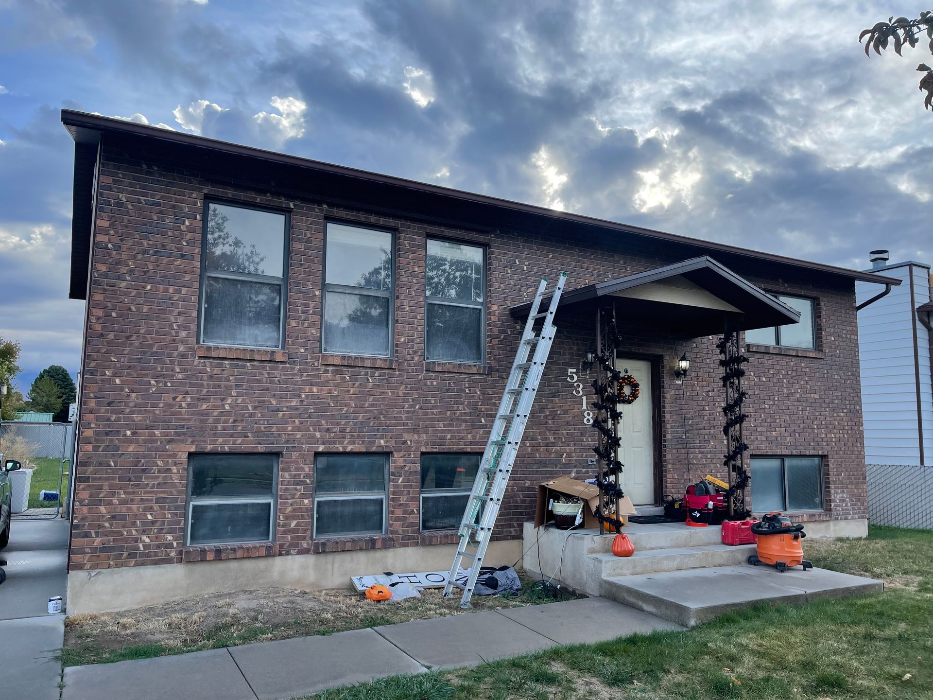 Brick house with ladder, porch, and tools on the lawn under a cloudy sky.