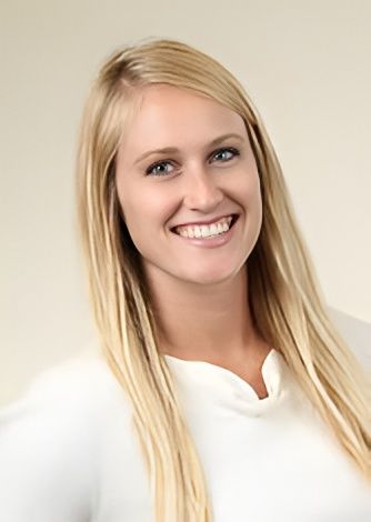 Blonde woman with blue eyes smiles. She wears a white sweater in front of a light background.