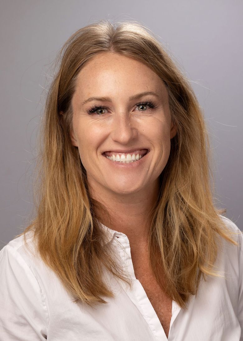 Blonde woman with blue eyes smiles. She wears a white sweater in front of a light background.