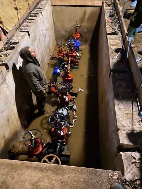 A man is standing in a muddy tunnel next to a row of valves.