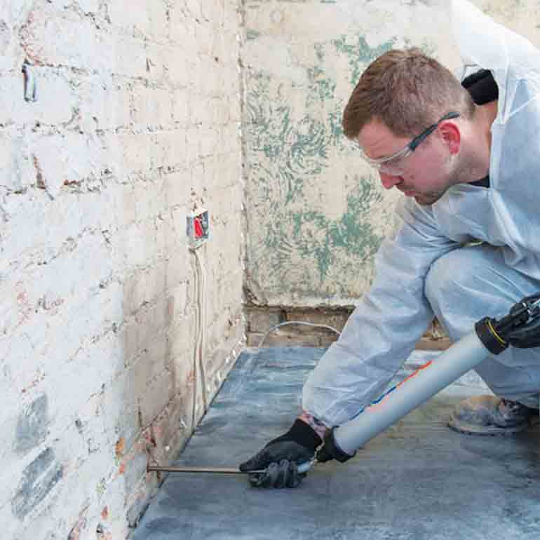 Man in protective suit injecting sealant into a brick wall base.