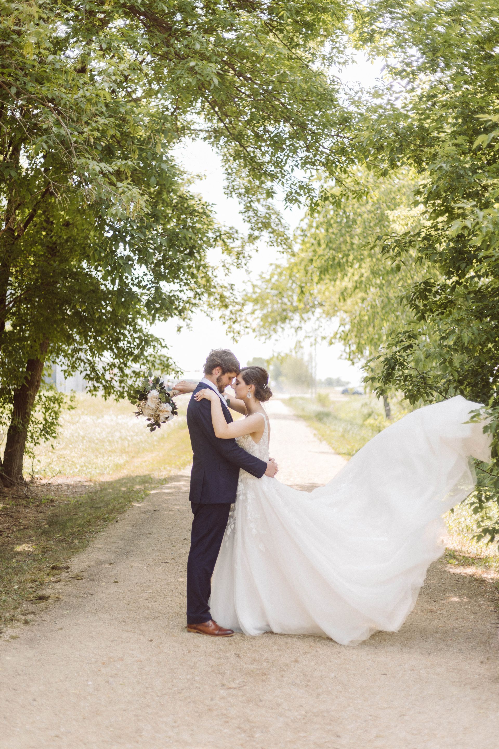 Groom in navy suit embraces bride in white gown on a tree-lined dirt road. Her train flows in the wind.