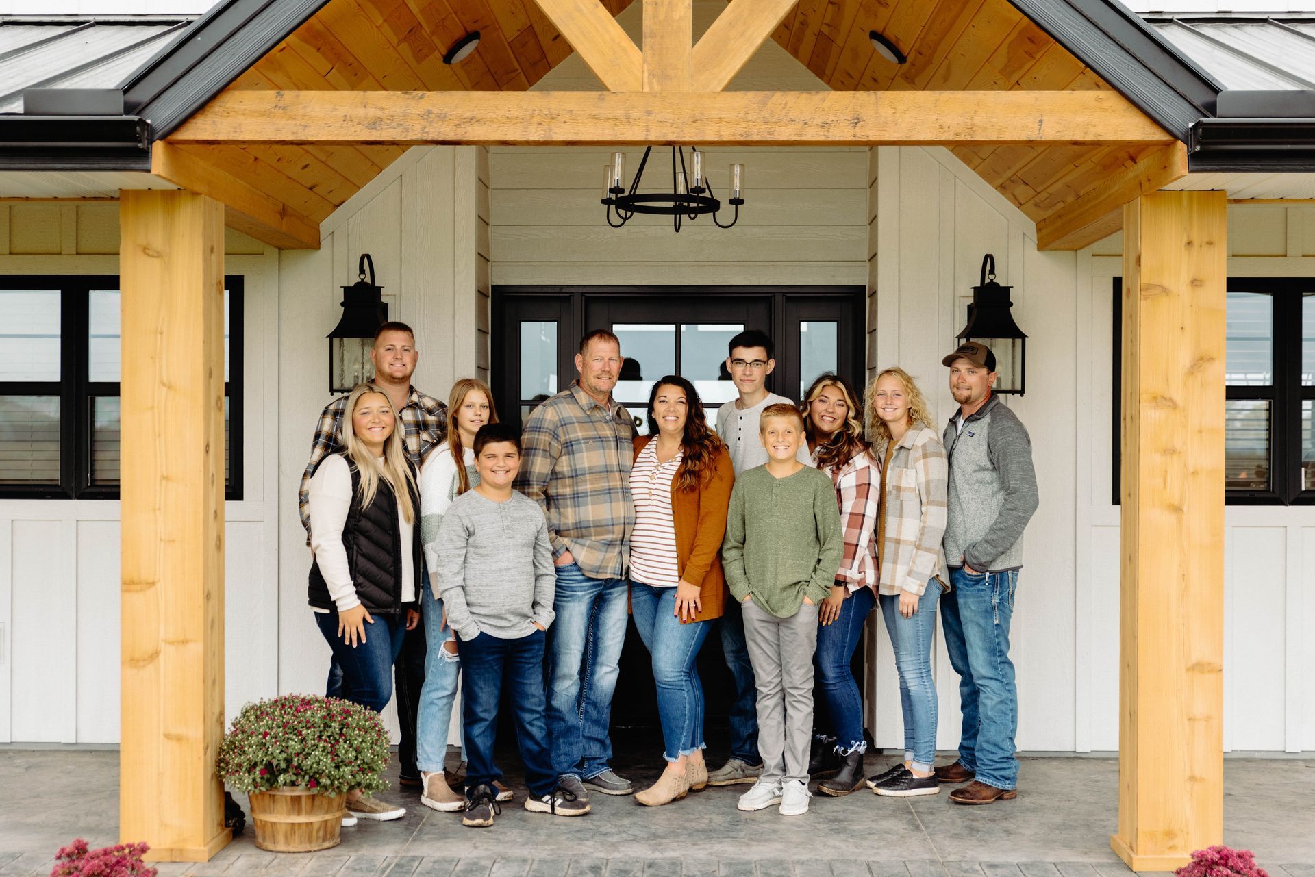 Family of 13 poses for a photo on porch; white farmhouse, wooden beams, casual attire, smiles.