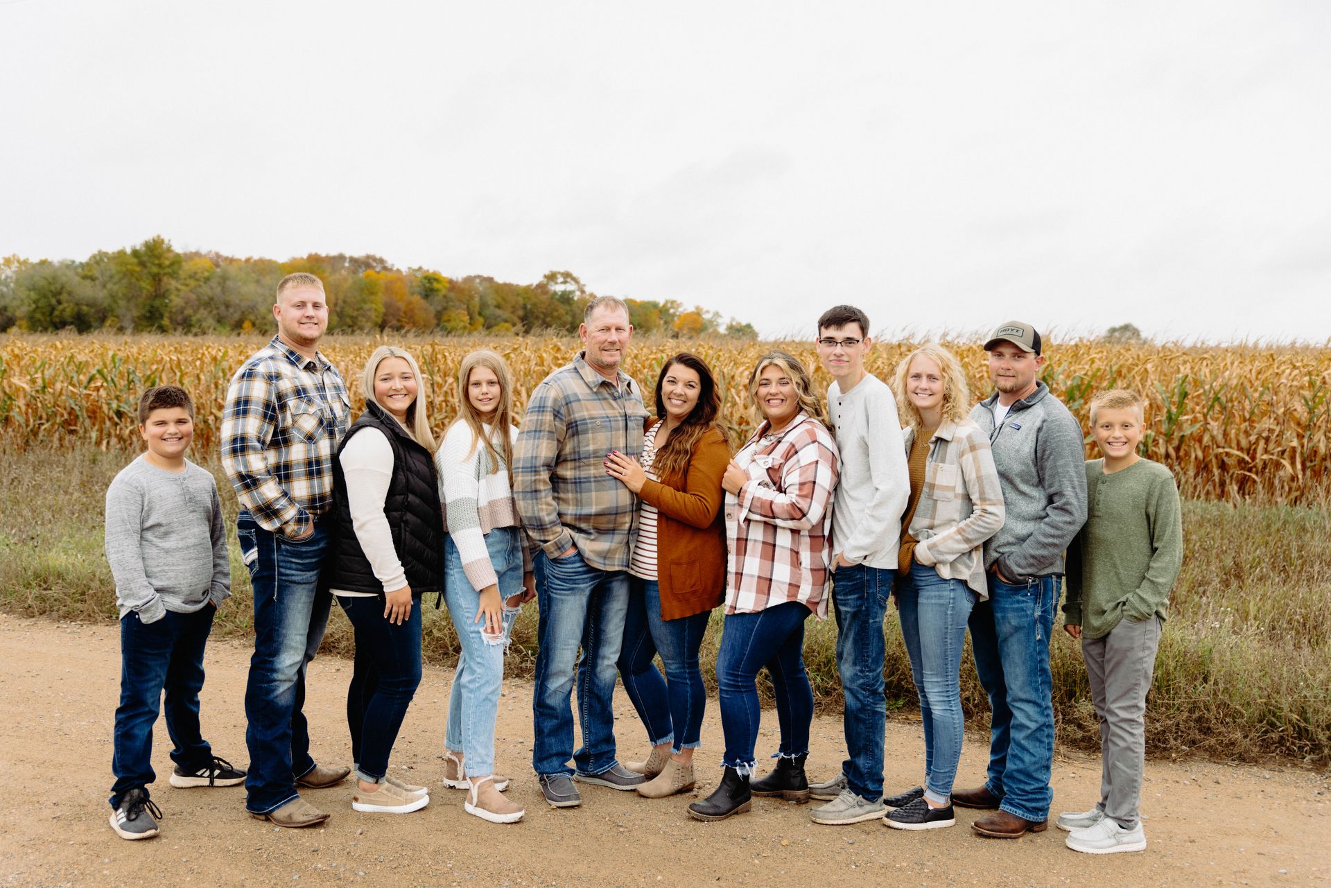 Family of 11 poses outdoors near a cornfield. They're wearing casual fall clothes and smiling.