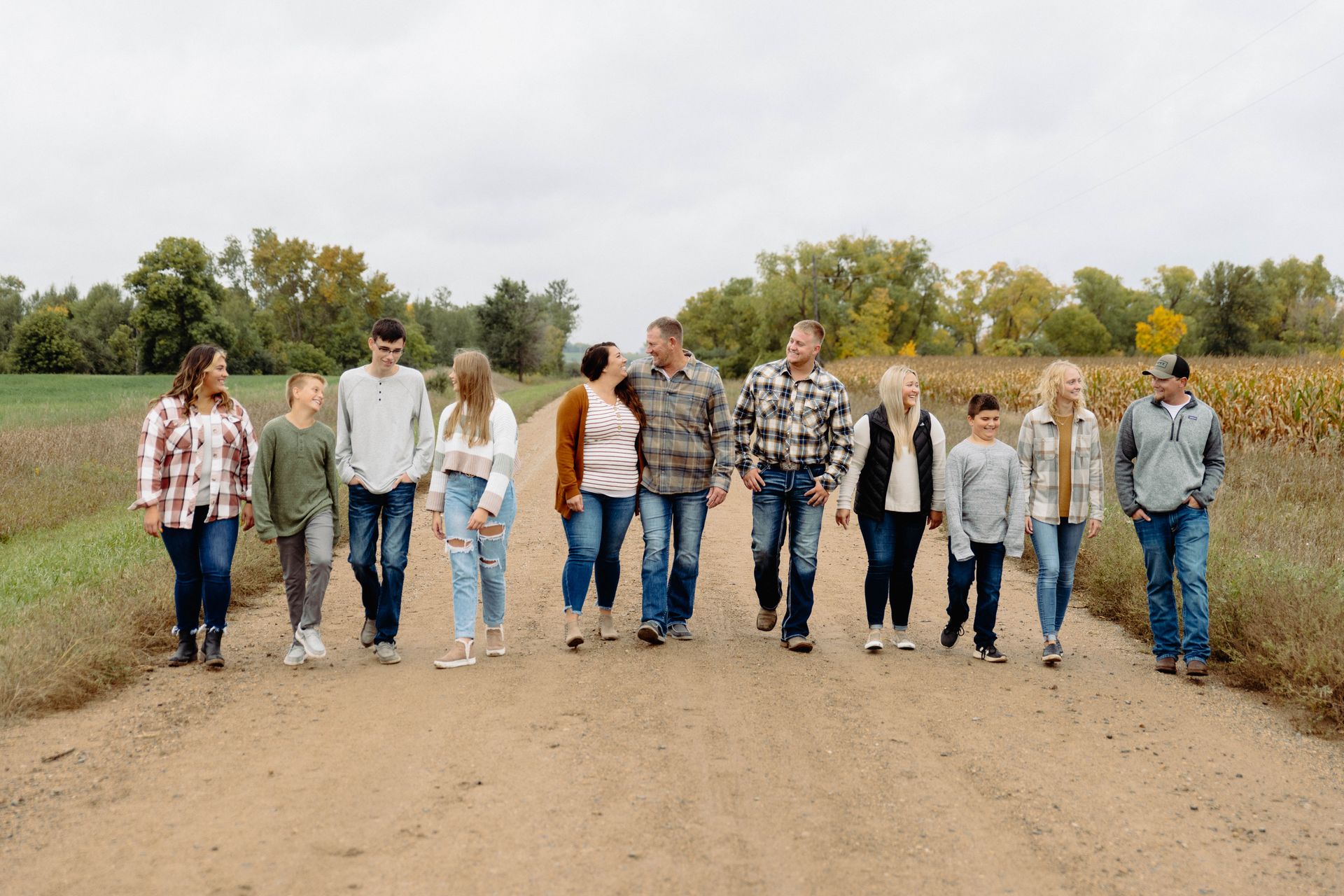 Family of 11 walking on a dirt road. Cloudy sky, green fields, and colorful trees in the background.