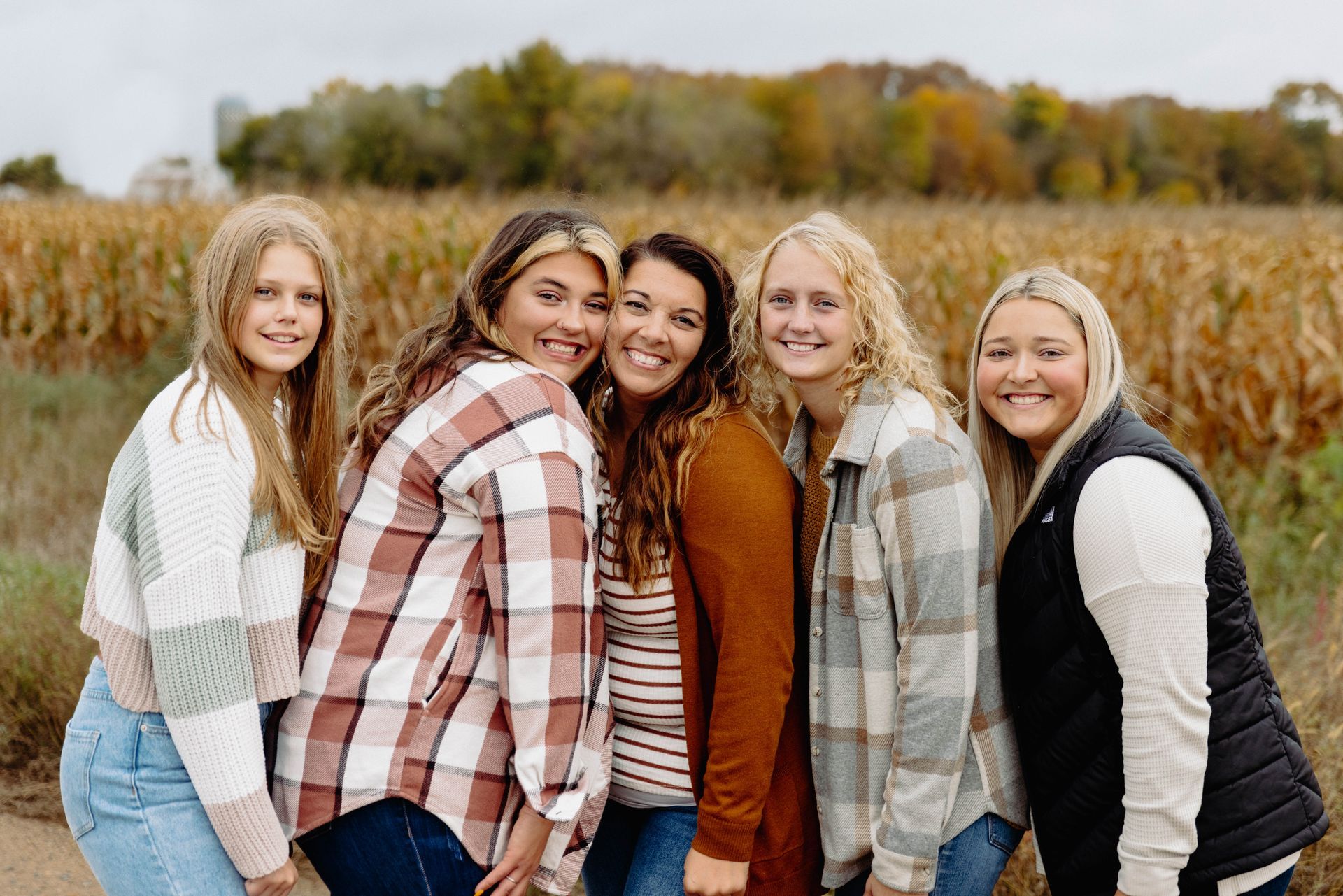 Five women smiling, hugging, in front of a cornfield. Wearing casual fall clothes, warm colors.