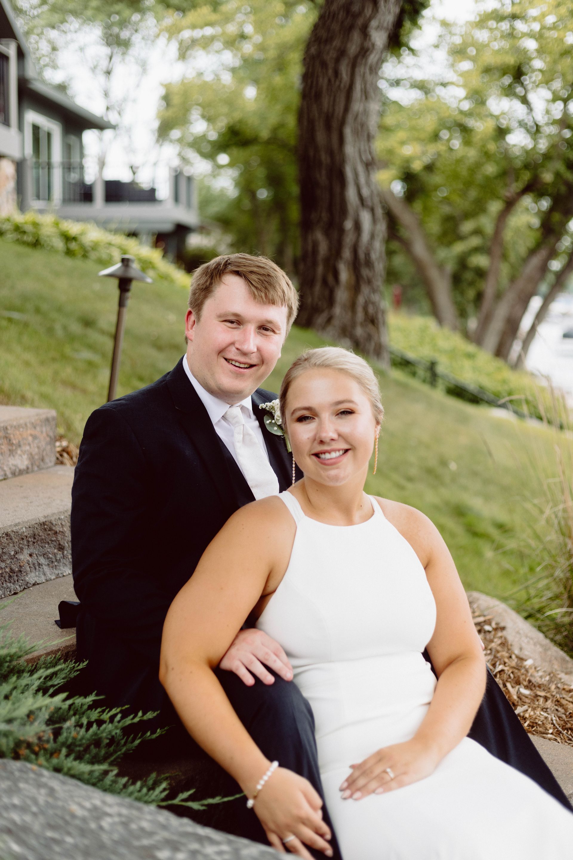 Minnesota Wedding Couple in wedding attire sitting on stone steps, smiling outdoors near a lake.