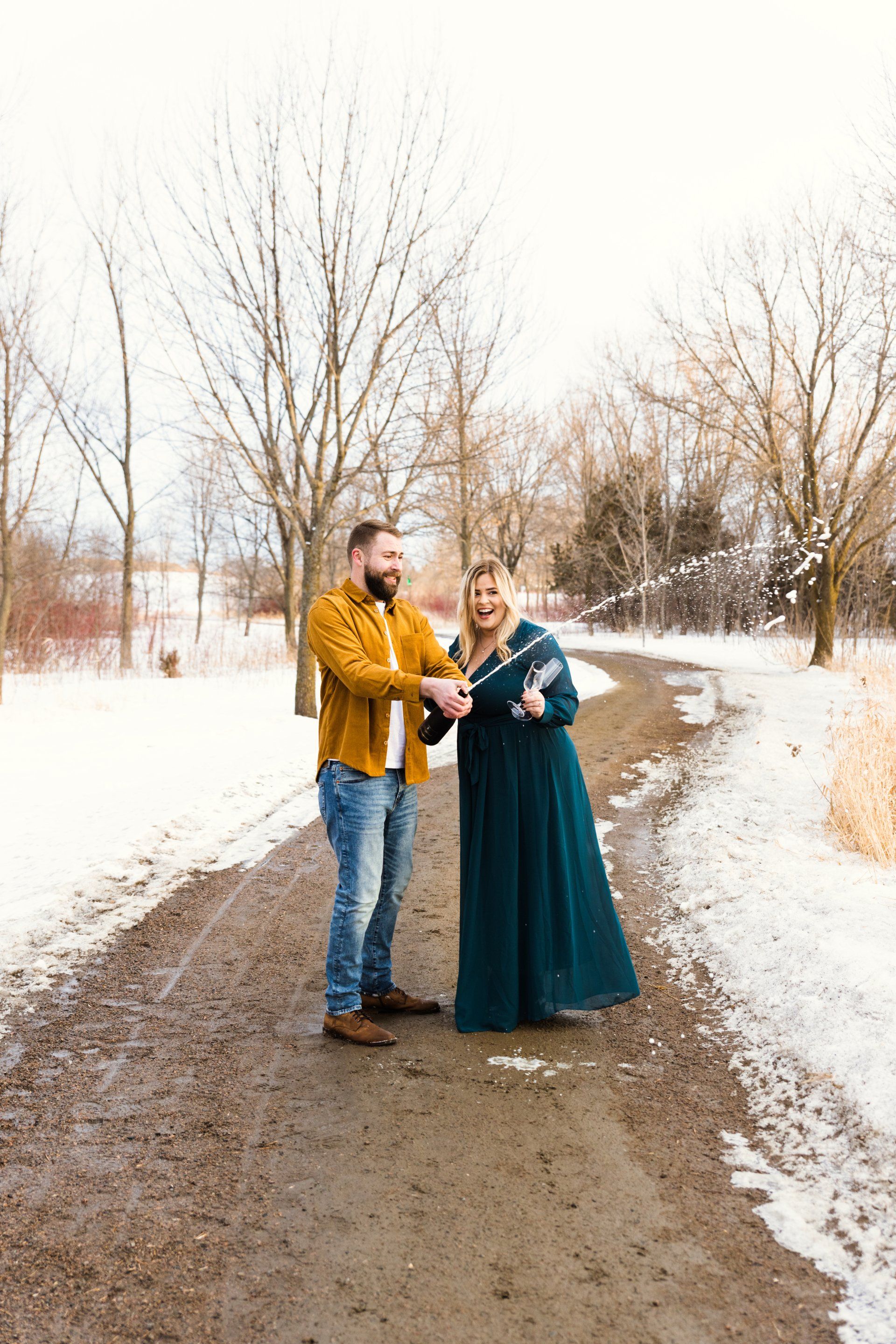 Couple celebrates with confetti in snowy path. Man in yellow jacket, woman in teal dress.