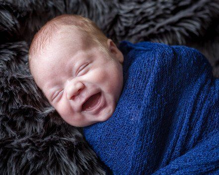 Smiling newborn baby wrapped in a blue blanket, lying on a dark, textured surface.