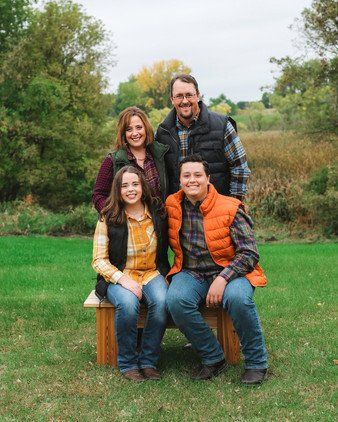 Family of four smiling, wearing vests, in a park. Two seated, two standing. Autumn colors, outdoors.