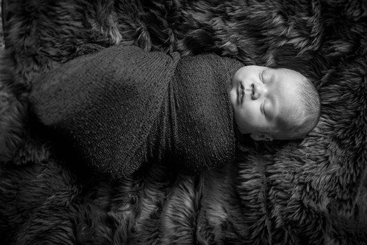 Newborn baby wrapped in dark fabric, sleeping peacefully on a furry brown surface.