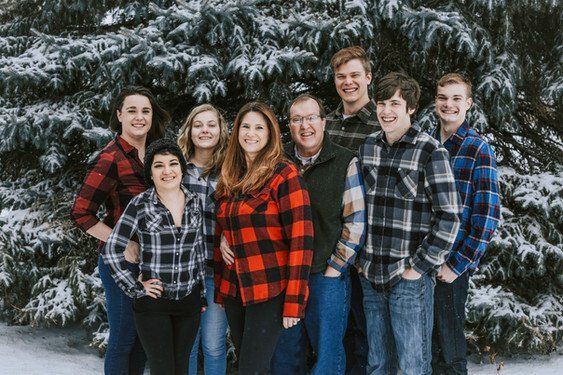 Family of nine poses in front of snowy evergreen tree. Wearing plaid shirts and smiling.