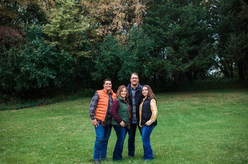 Family of four standing in a grassy field with trees behind them. They are wearing vests and jeans.