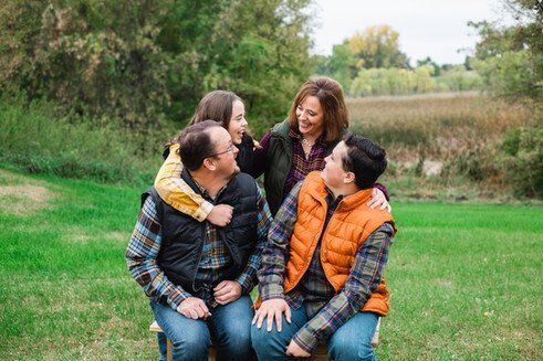 Family of four smiling outdoors; mother hugs daughter; father and son look at each other.