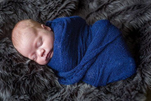 Newborn baby swaddled in blue blanket, sleeping on a gray, furry blanket.
