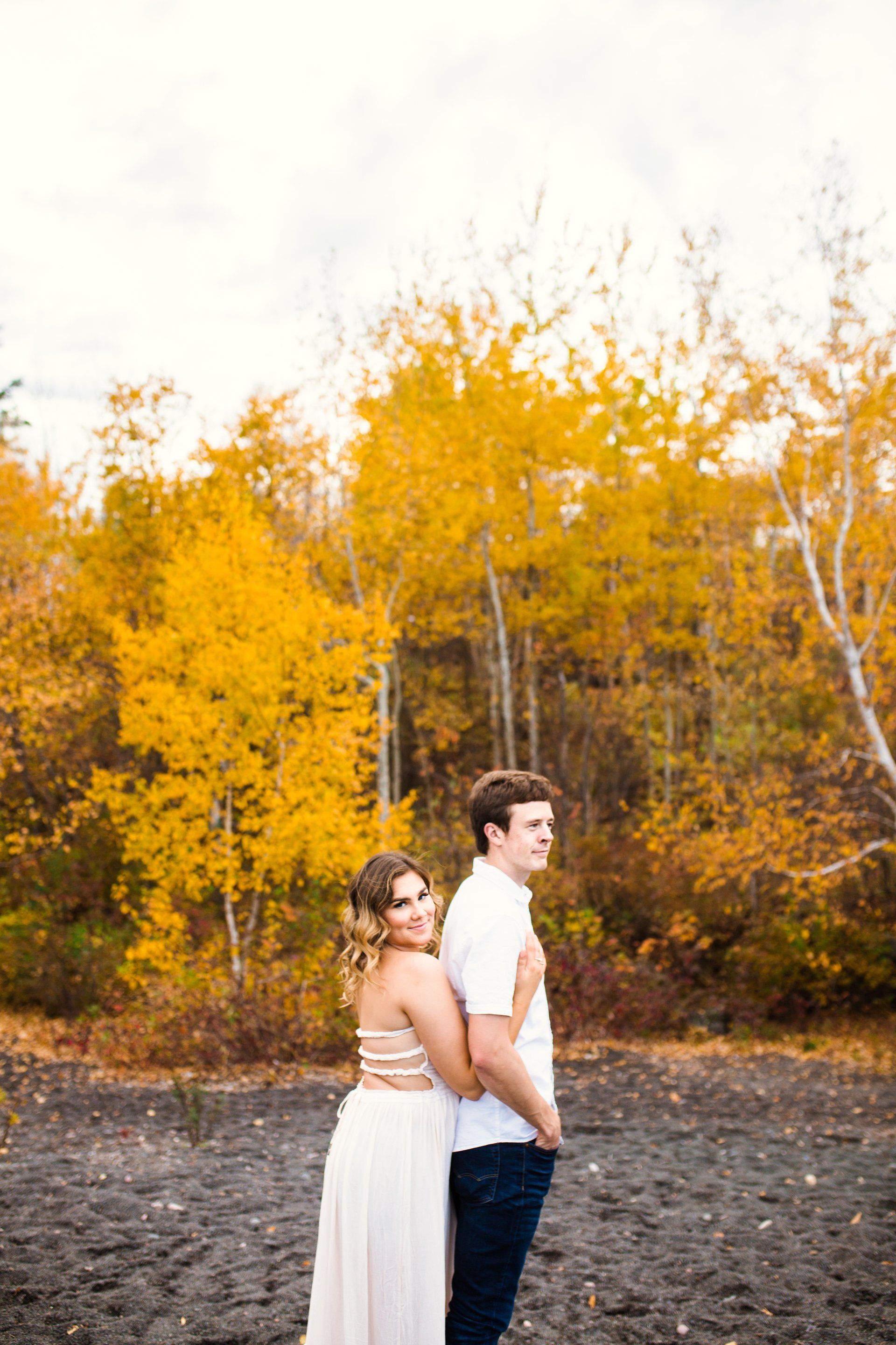 Couple in white dress and shirt embrace, standing before fall foliage.