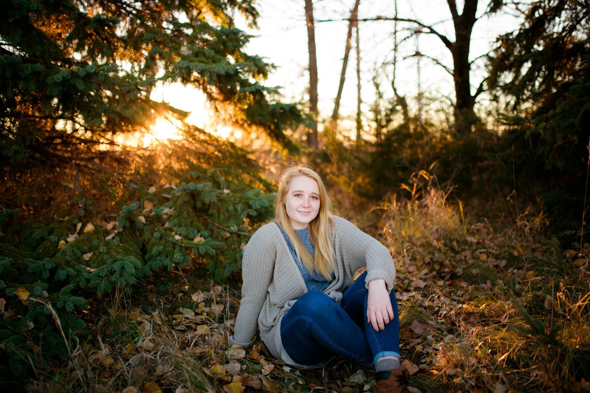 Blonde woman sitting outside in fall foliage, smiling at the camera. Golden sunlight peeks through trees.