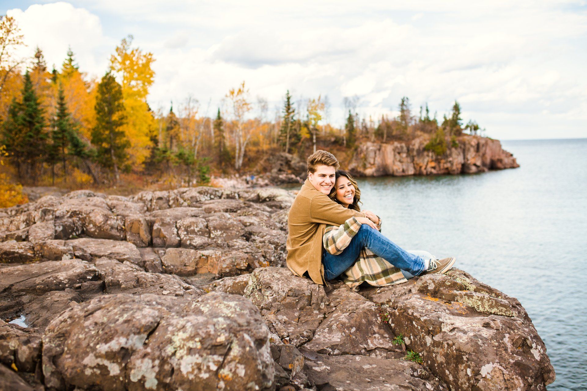 Couple hugging on rocky cliff overlooking lake with fall foliage.