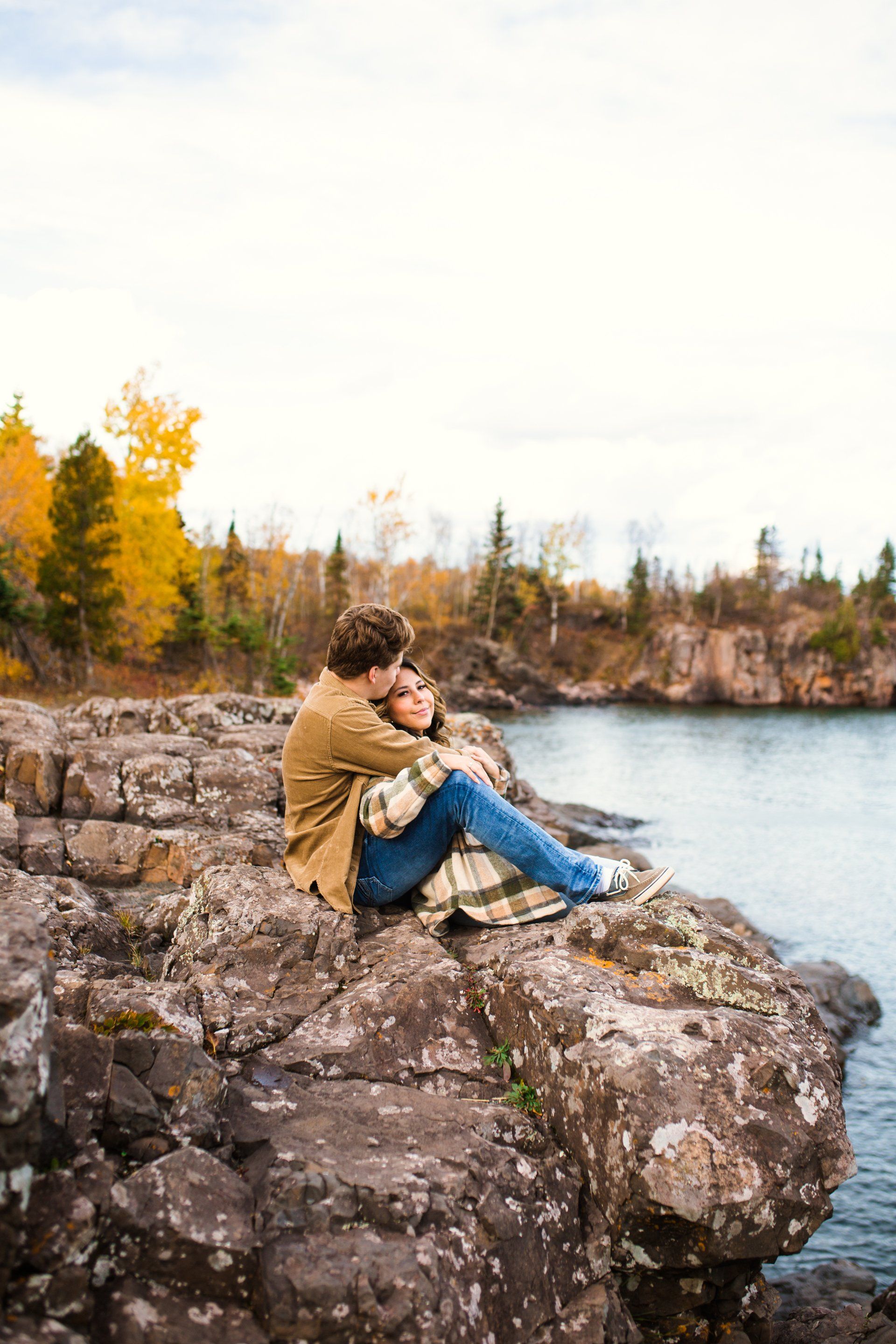 Couple embraces on rocky cliff overlooking water, fall foliage in background.