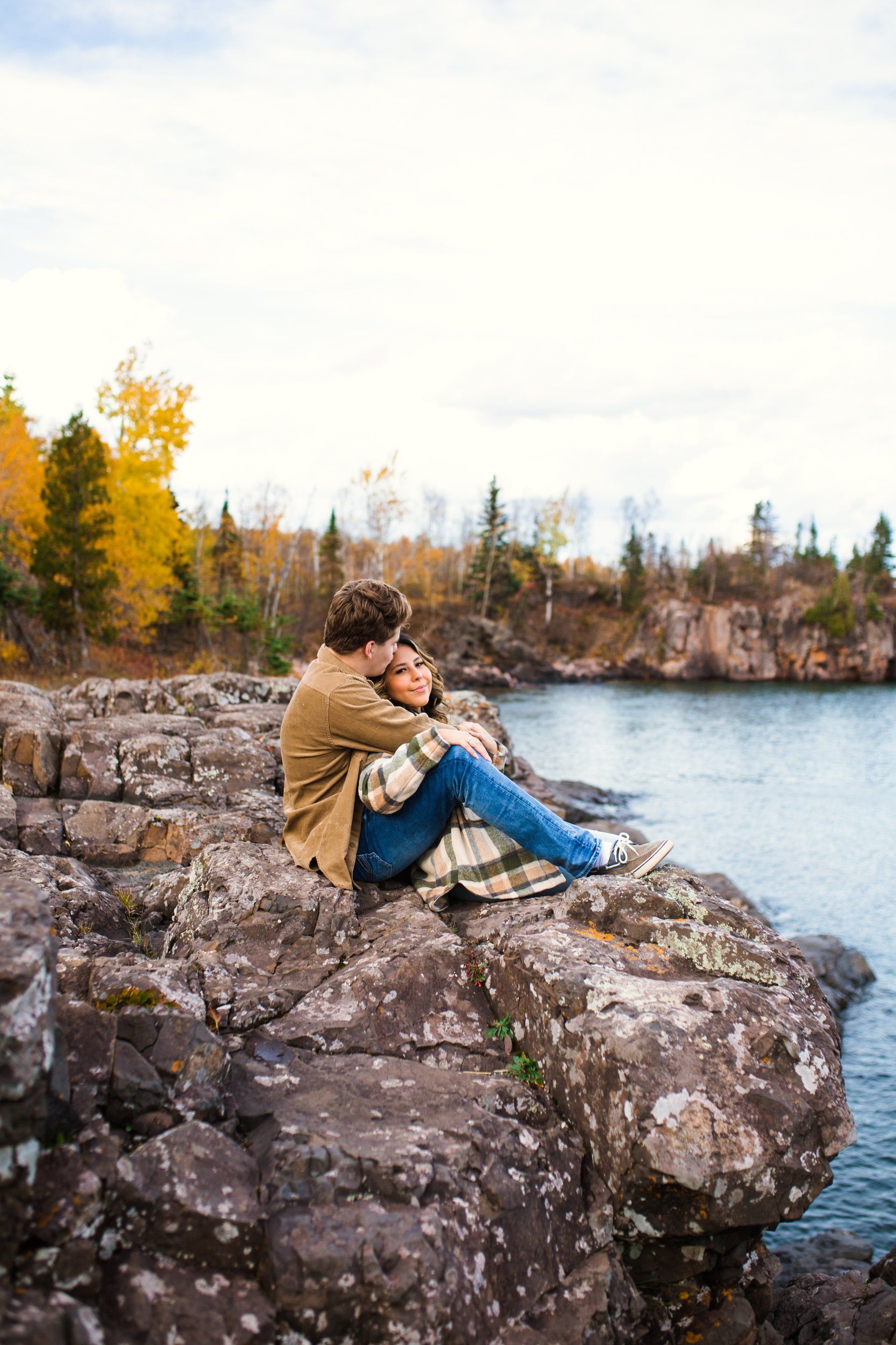 Couple embraces on a rocky cliff overlooking a lake with autumn foliage in the background.