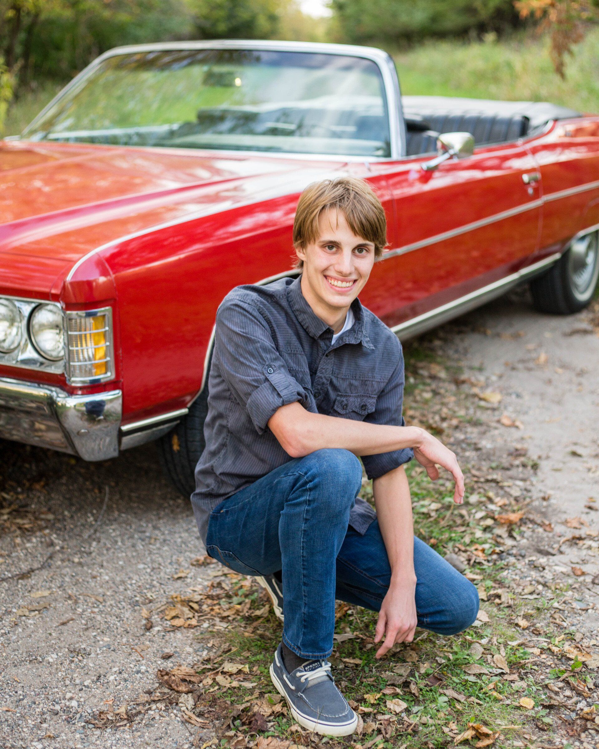 Young man kneels next to a red convertible car; he smiles, wearing a gray shirt, blue jeans, and dark shoes.