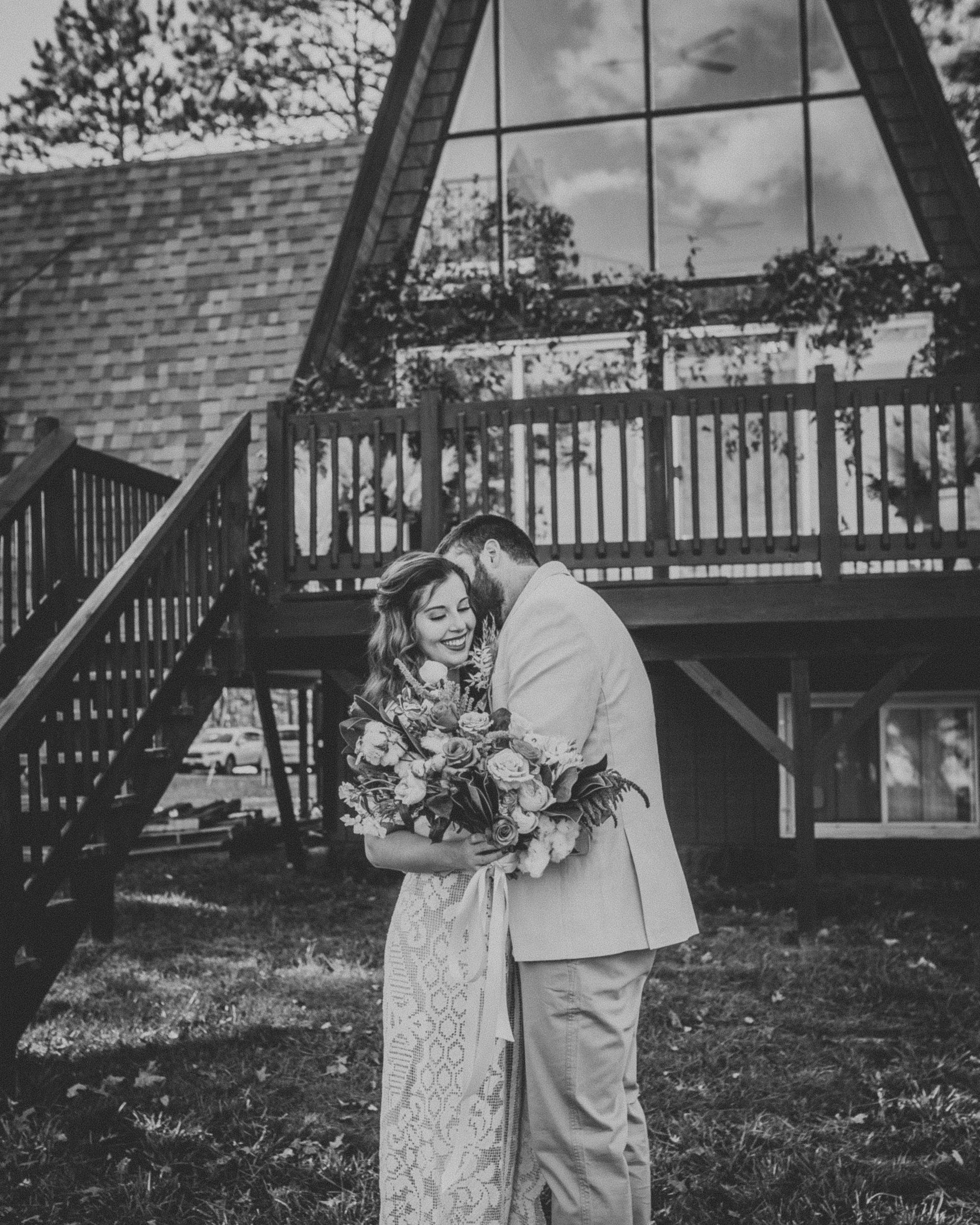 Bride and groom embrace outside A-frame house; bride holds bouquet, groom whispers; black and white.