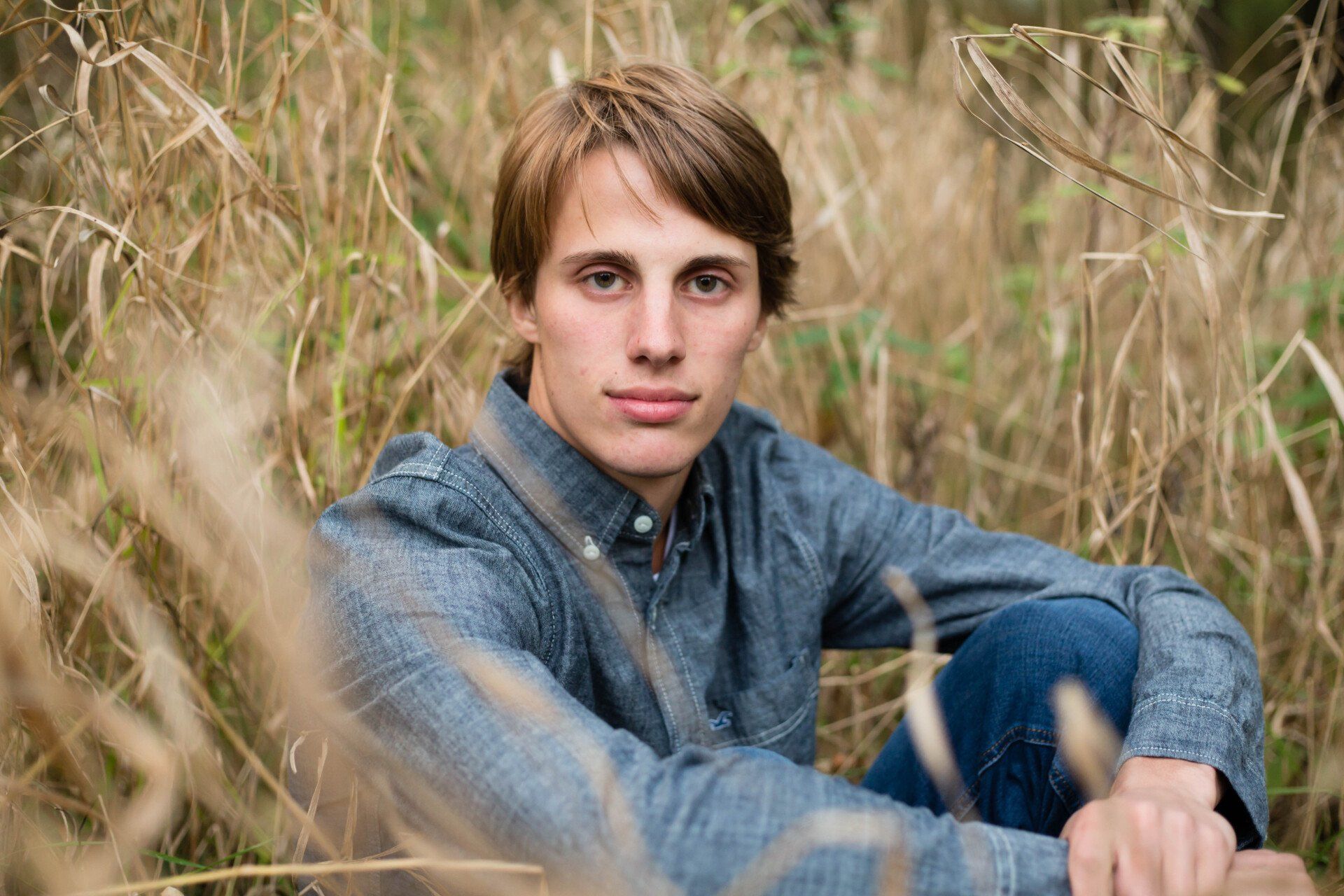 Young man with light brown hair, blue denim shirt, and jeans, seated in tall dry grass.