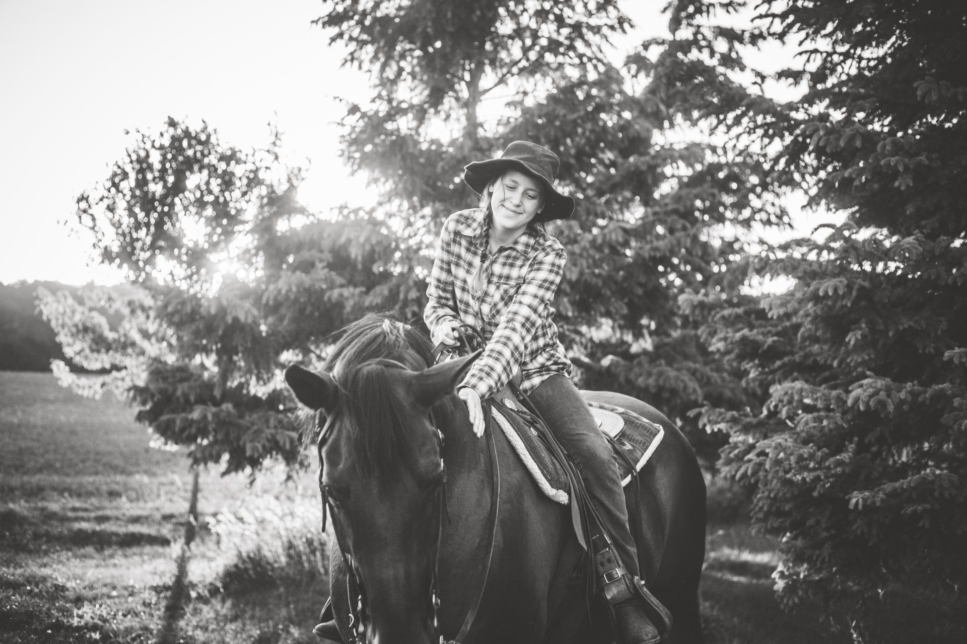Cowgirl smiles while riding a horse in a field, wearing a plaid shirt and hat. Black and white.