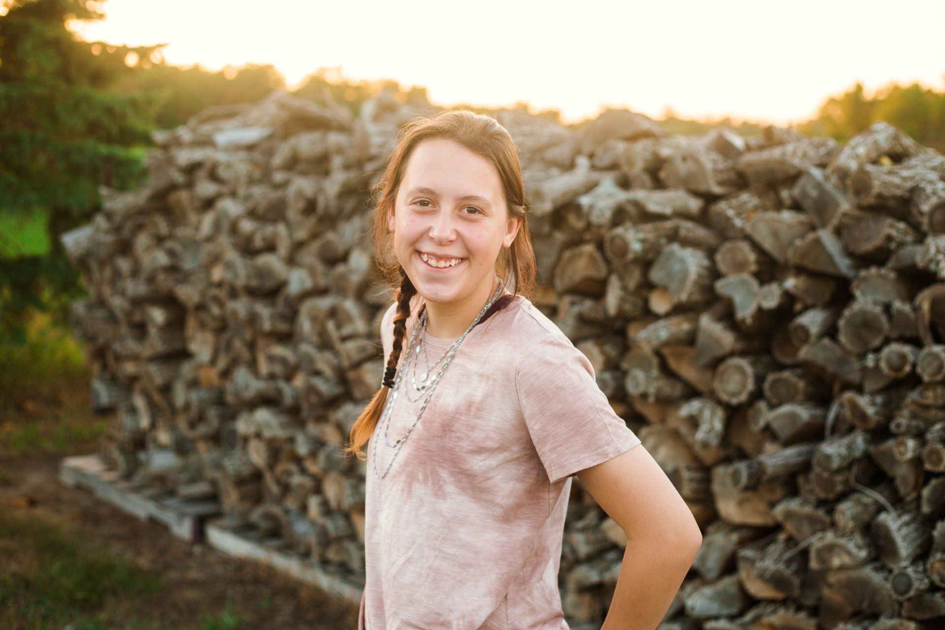 Smiling girl with a braid, in front of a pile of firewood, wearing a tie-dye shirt.
