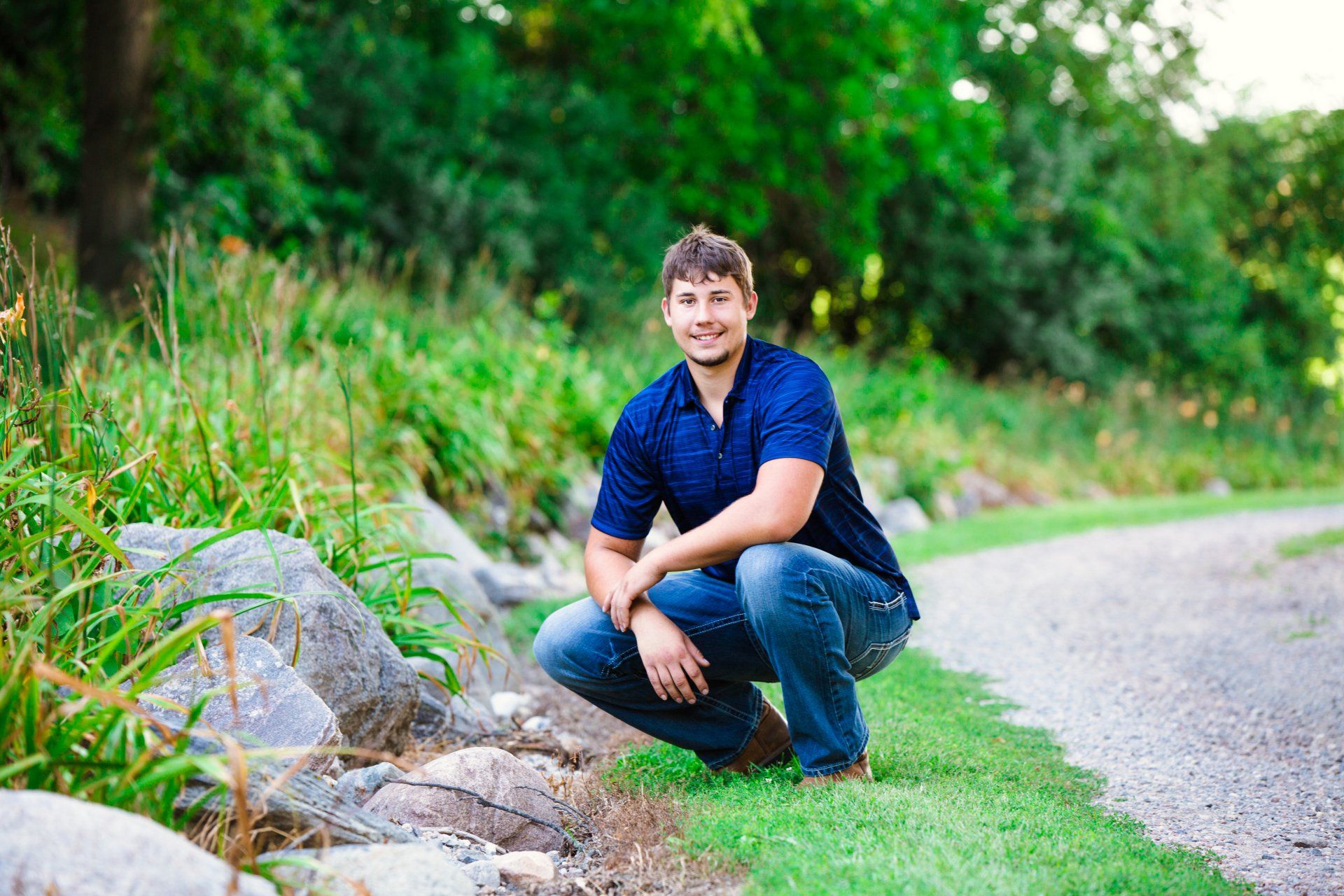 Young man in blue shirt and jeans squats on a path near rocks and greenery, smiling at the camera.