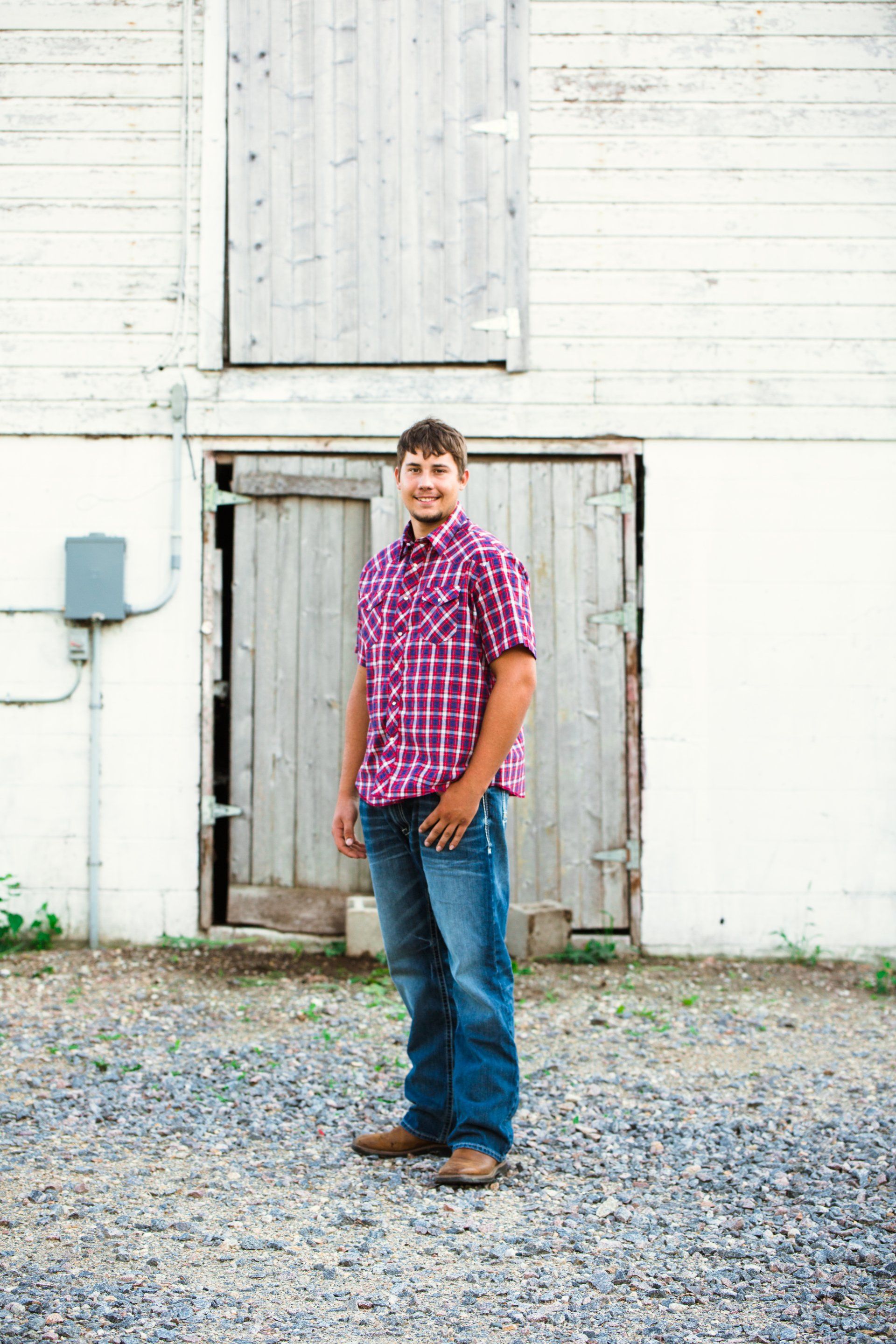 Young man stands in front of a weathered white barn, wearing a plaid shirt, jeans, and boots.