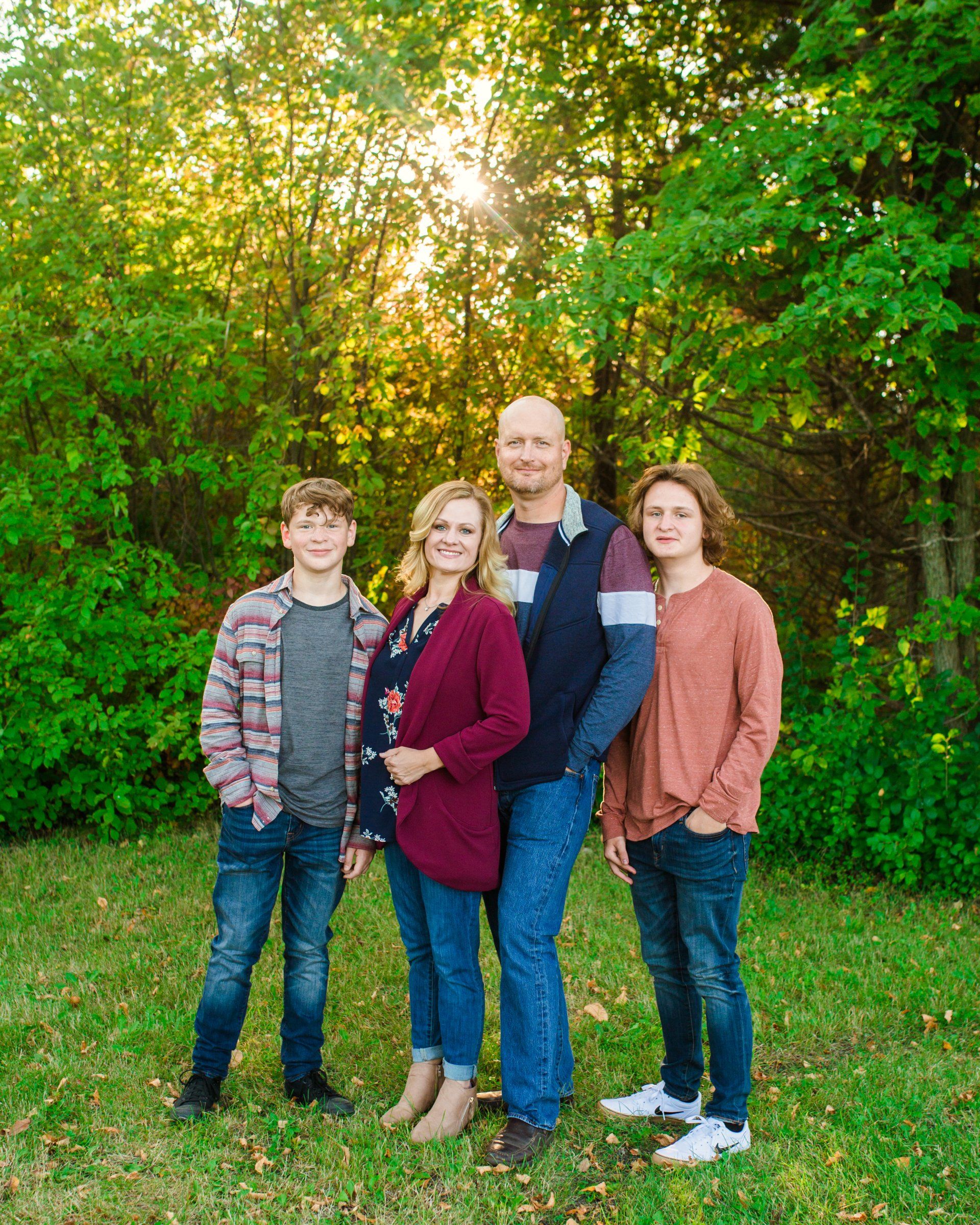 Family of four standing in a grassy area with trees, smiling at the camera; warm sunlight.
