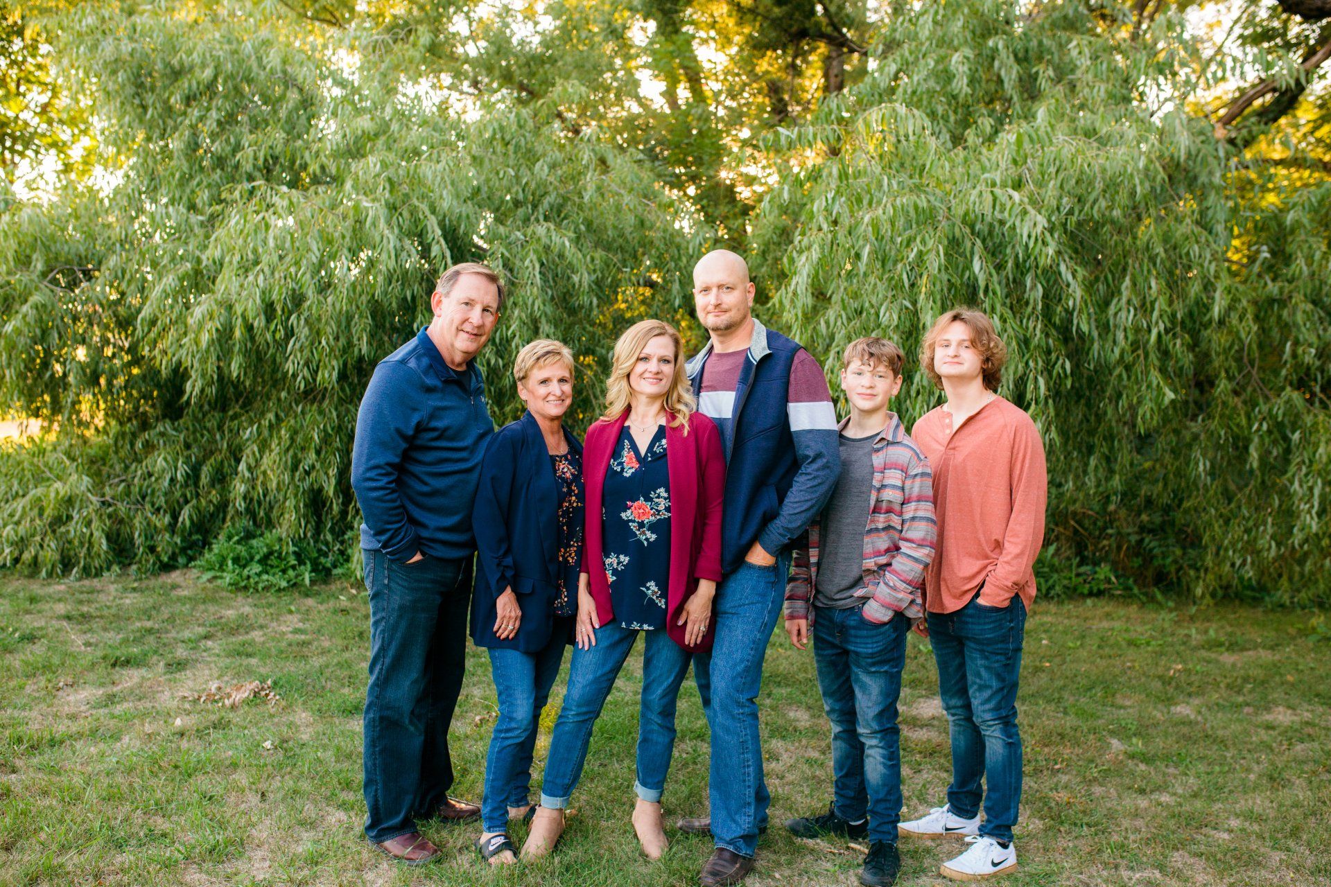 Family portrait, six people smiling outside on grass; green trees in the background.