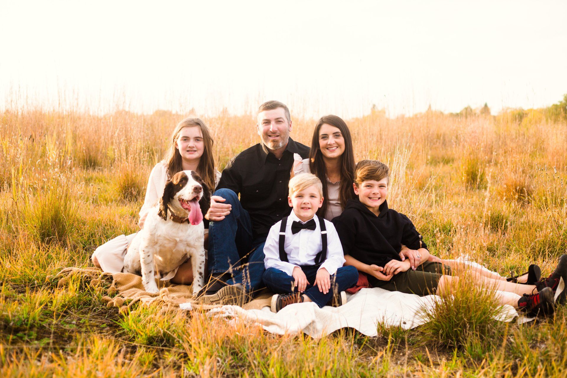 Family of five and dog in tall grassy field, seated on blanket, golden light.