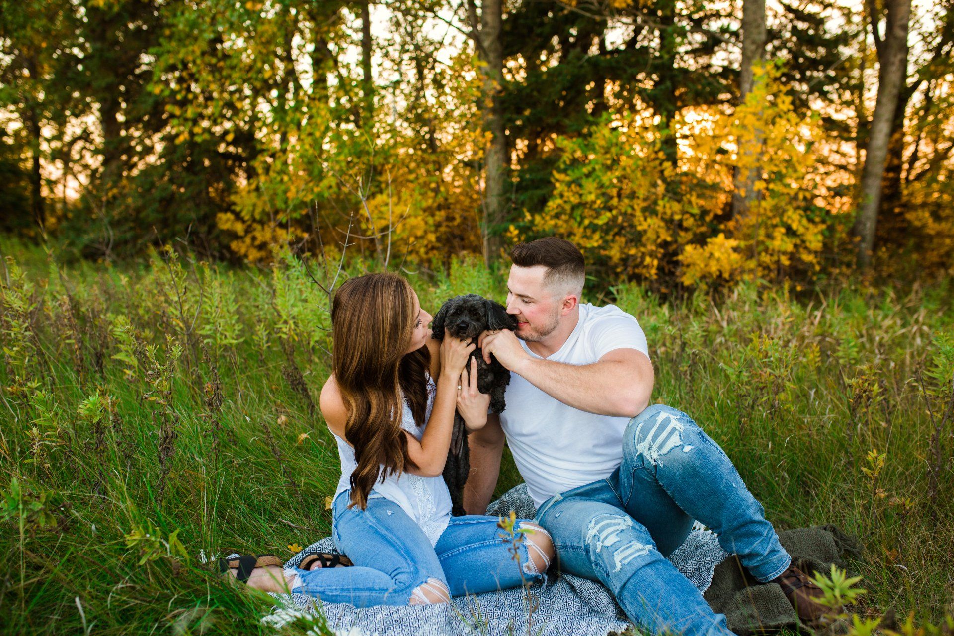 Couple and dog sitting in a grassy field, embracing. Woman kisses the dog while the man holds it.