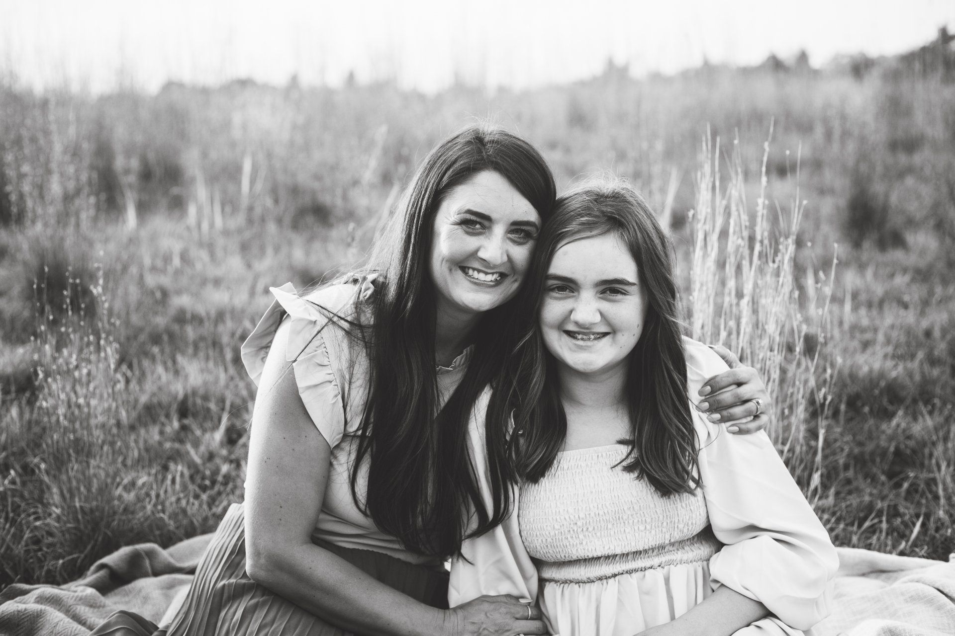 Woman with long hair and girl in dress smile, embrace outside.