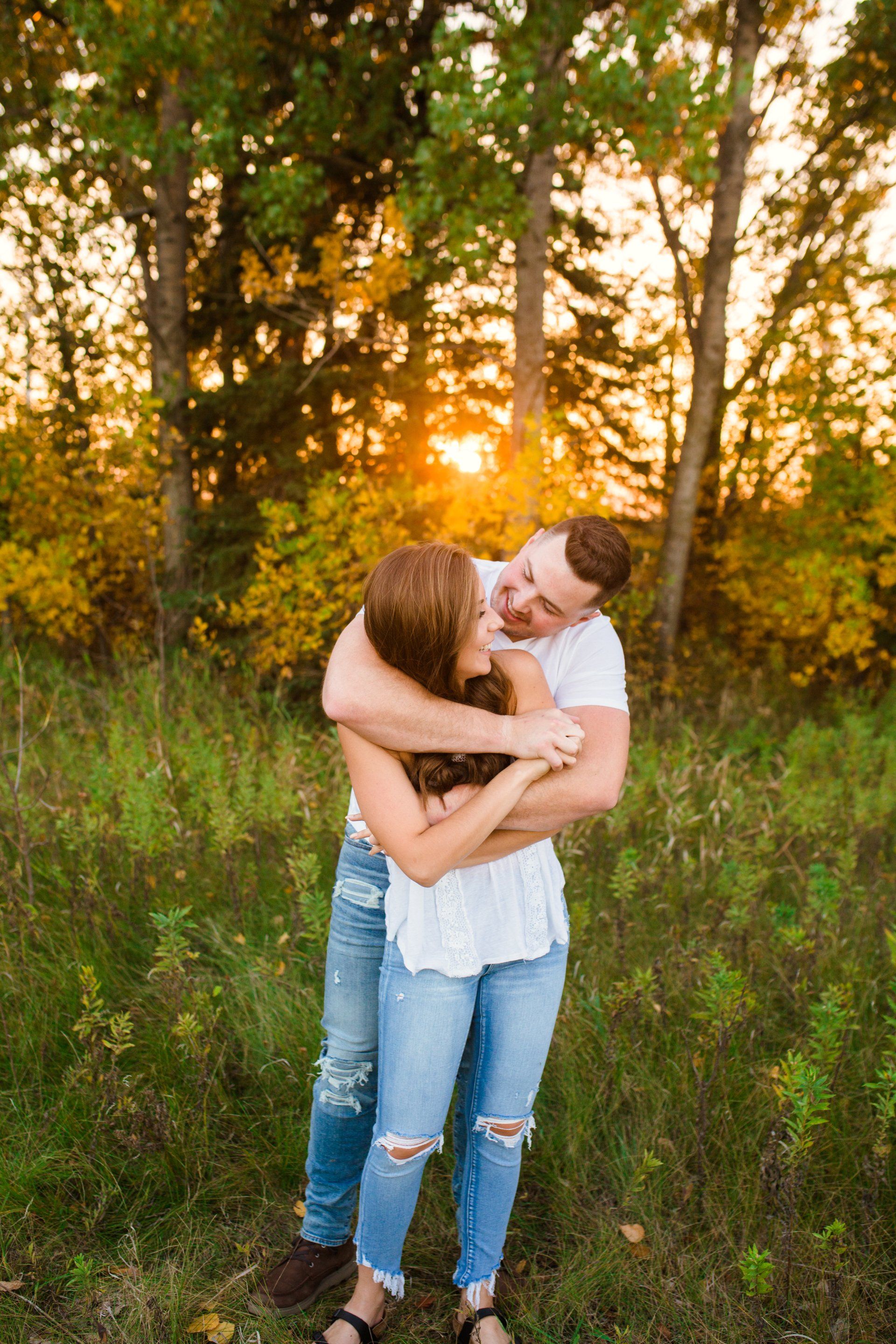 Couple embraces in field; sunset glows behind trees. Man hugs woman, looking into her eyes.