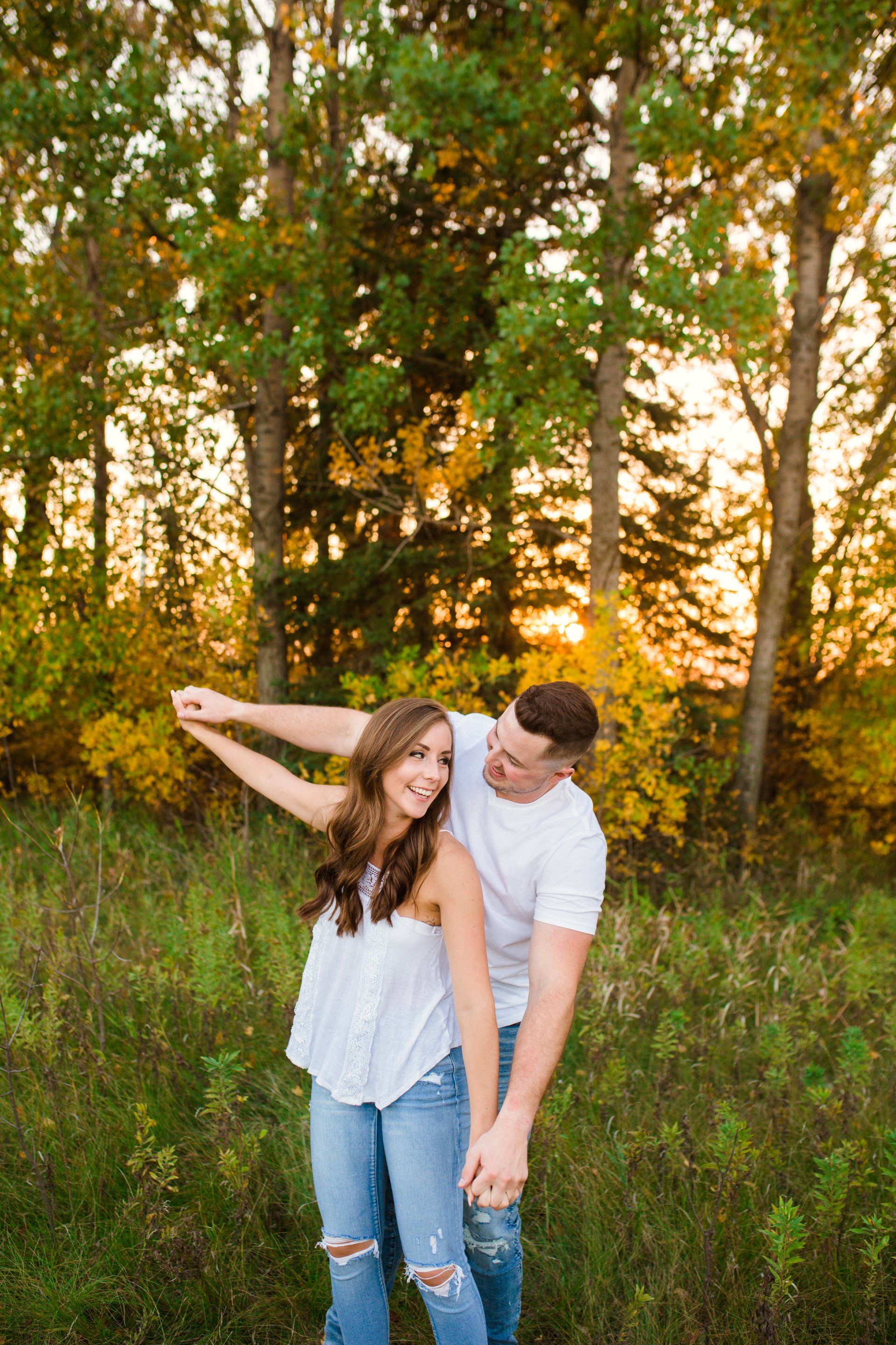 Couple in field, man embracing woman, smiling. Golden sunlight, trees in background.