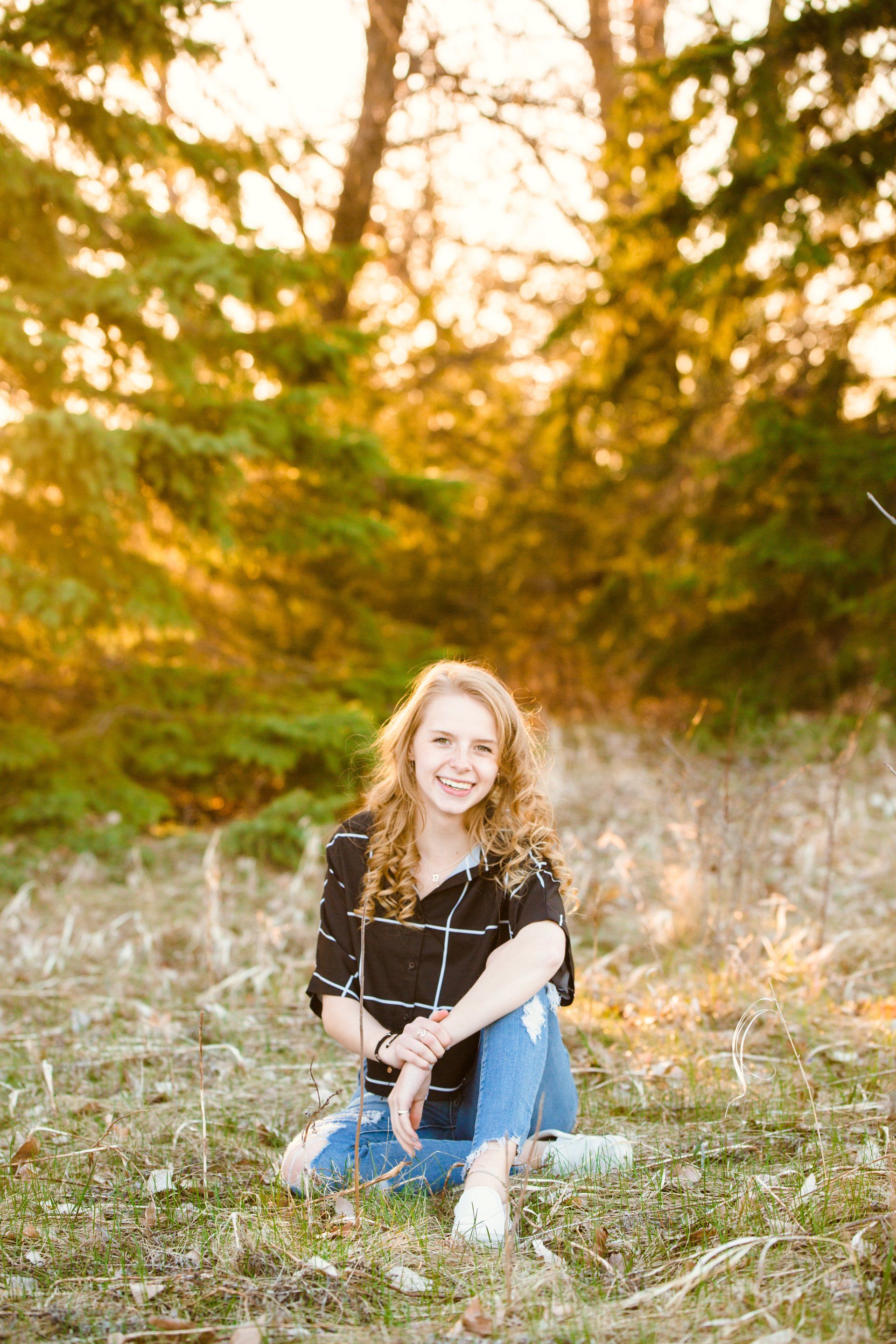 Woman with curly hair smiles, sitting outside, wearing a plaid top and ripped jeans.