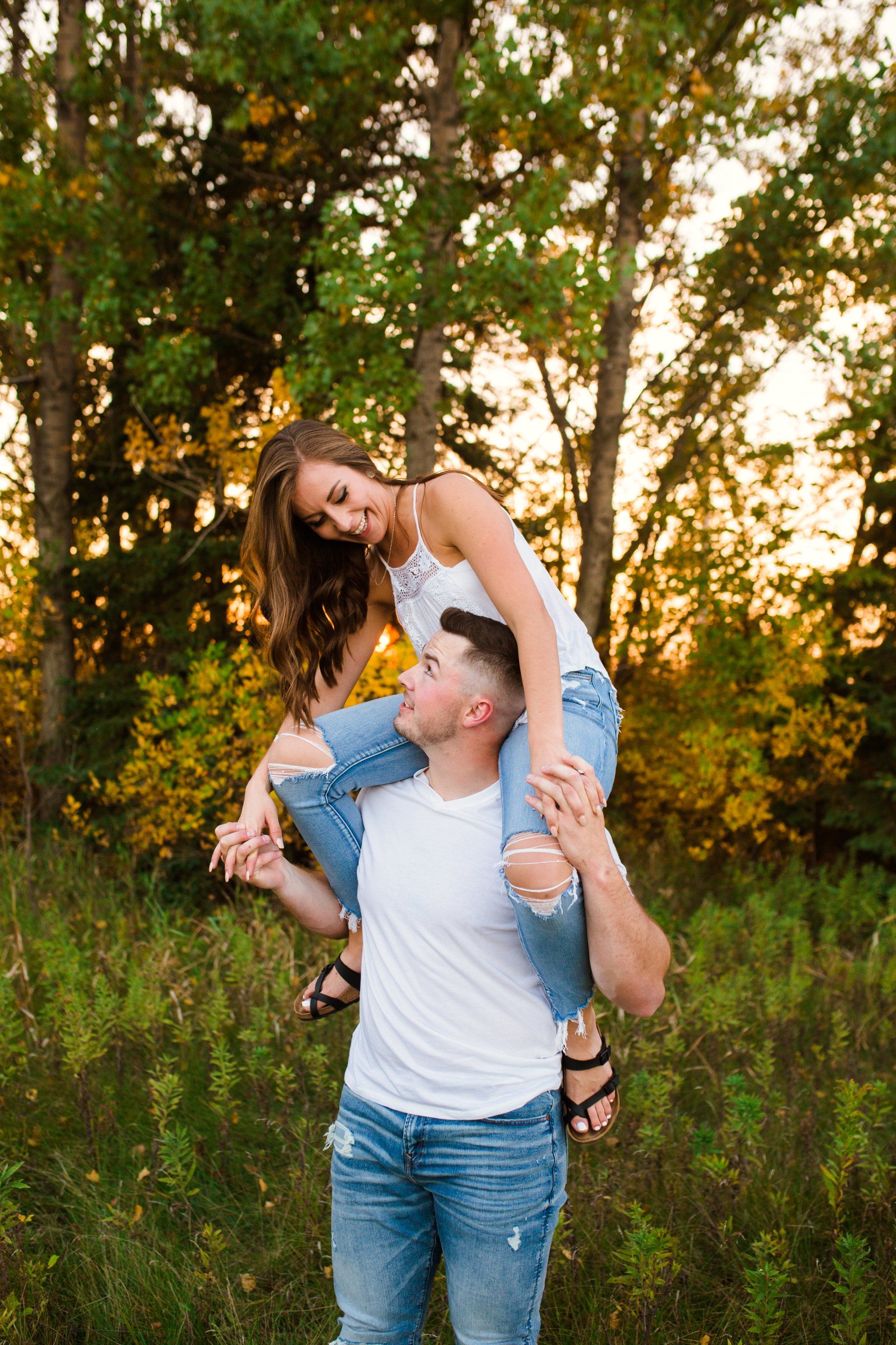 Couple in ripped jeans embrace; woman on man's shoulders in a field at sunset.