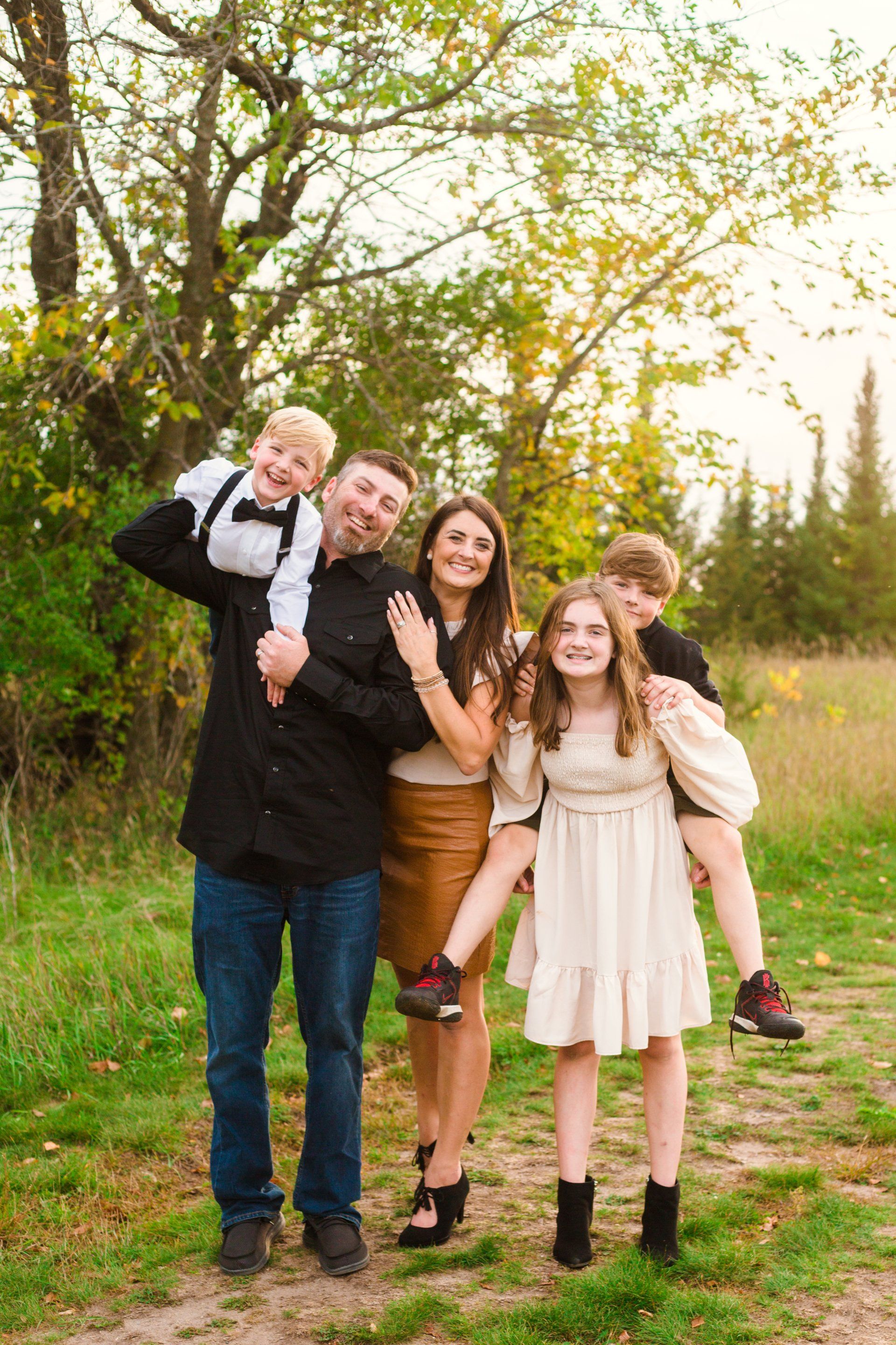 Family of five smiling outdoors; dad holding child on shoulders, another child piggybacking.