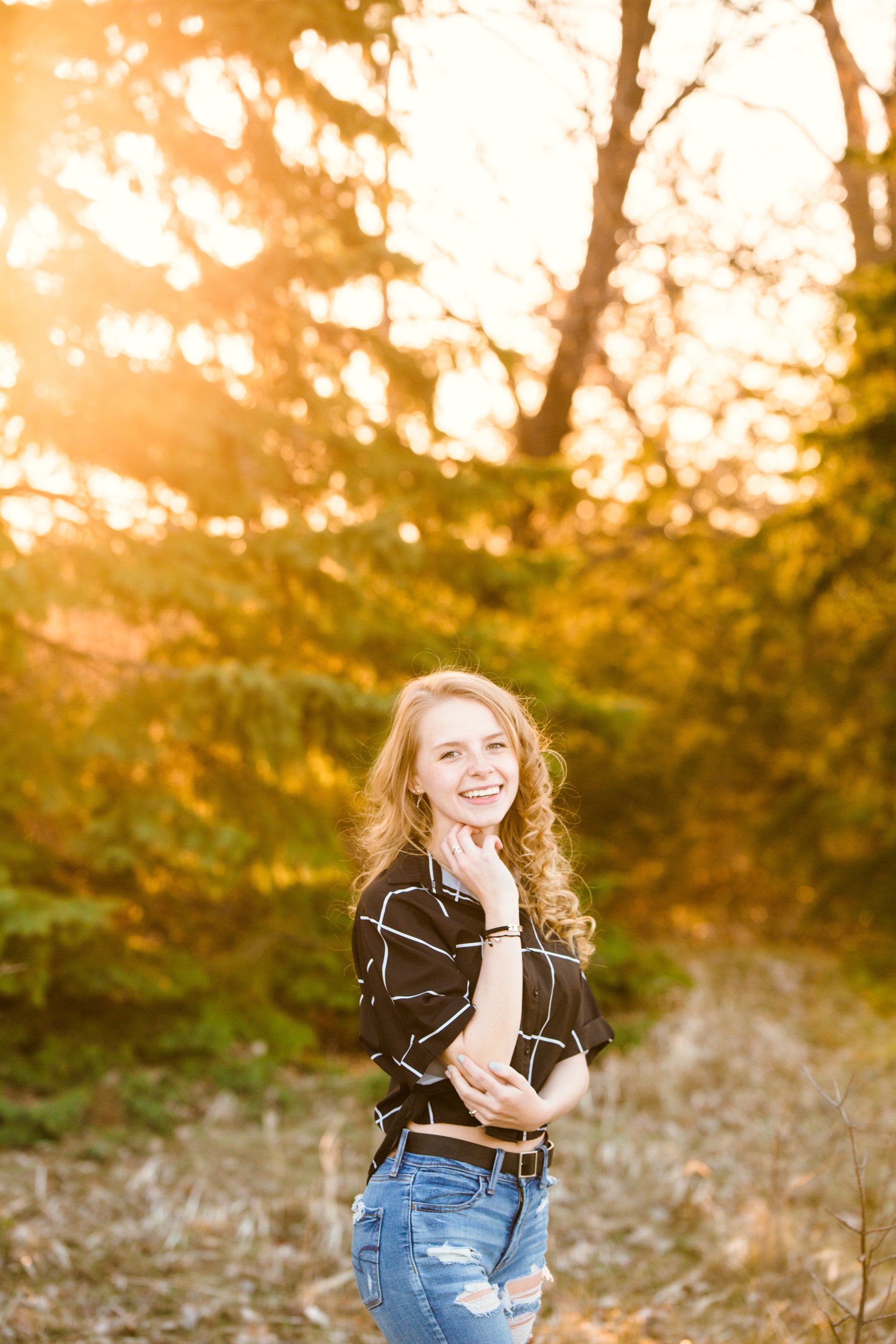 Woman with curly hair smiles in a forest at sunset.