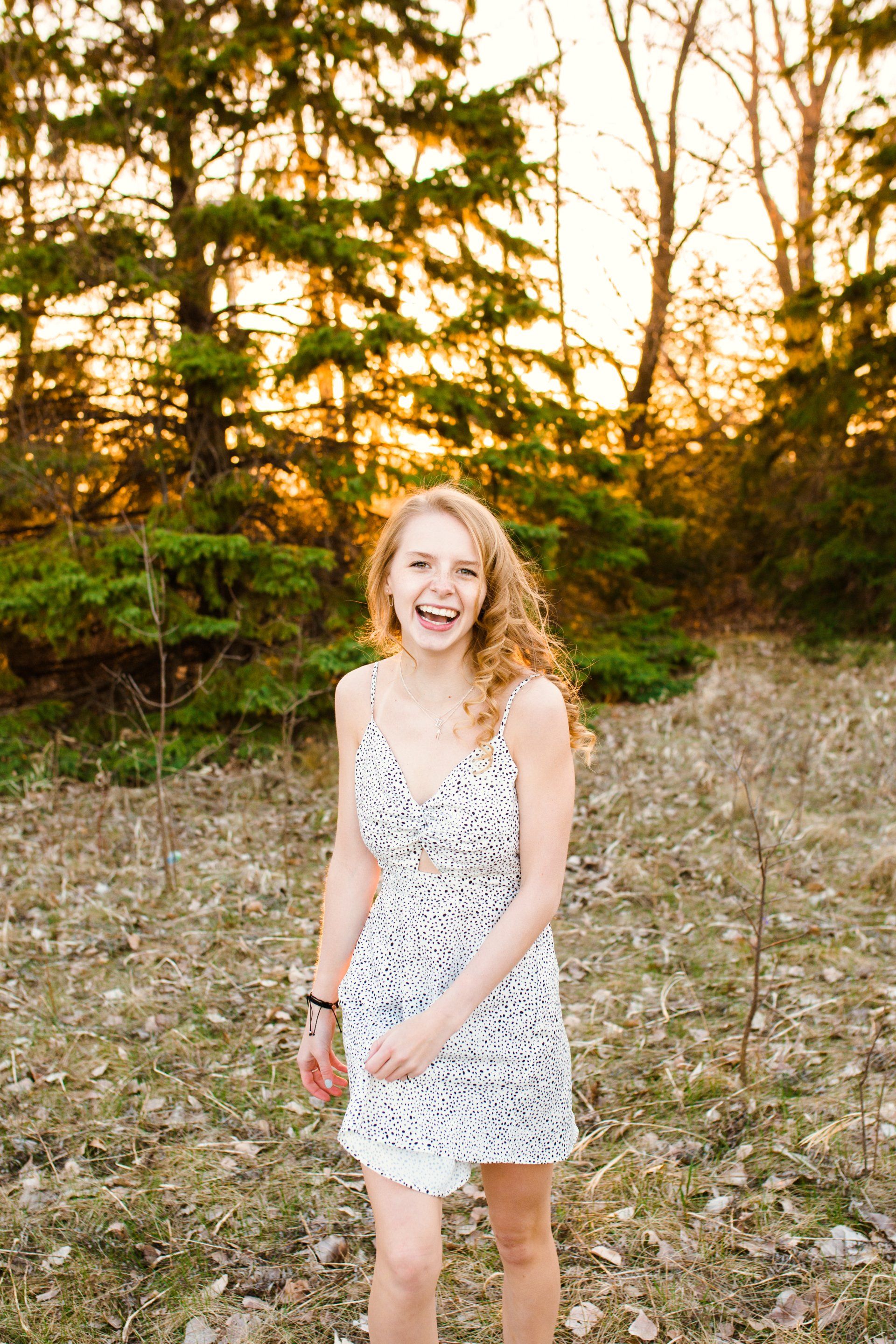 Woman with curly hair laughs joyfully in a white dress with a forest backdrop at sunset.